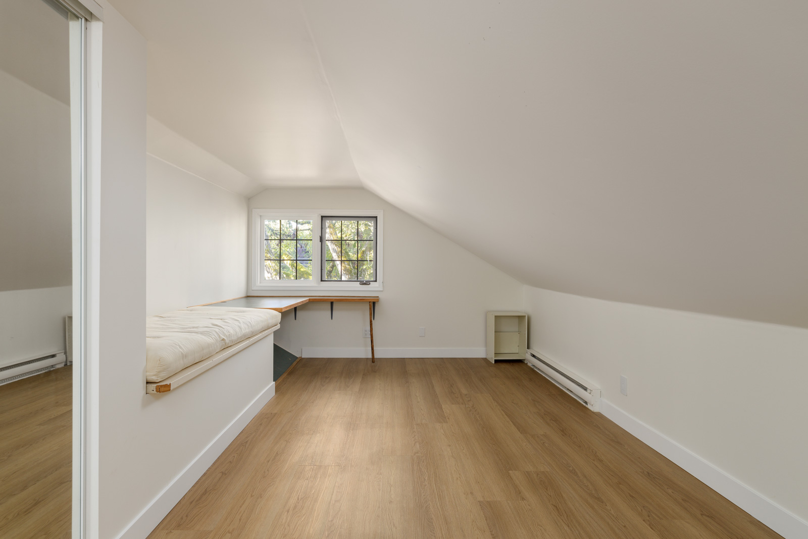 An empty attic room with sloped ceiling, wooden floor, a small desk beneath the window, and a built-in bench with a cushion on the left.