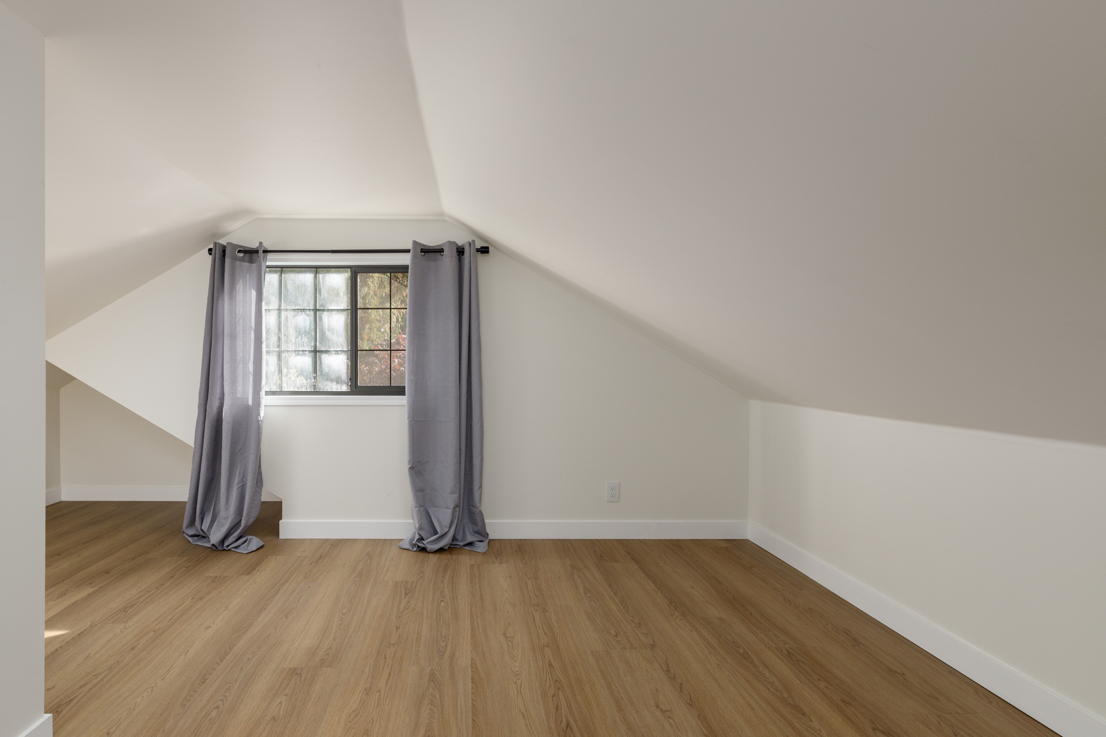 An empty attic room with sloped ceilings, a window with gray curtains, and light wood flooring.