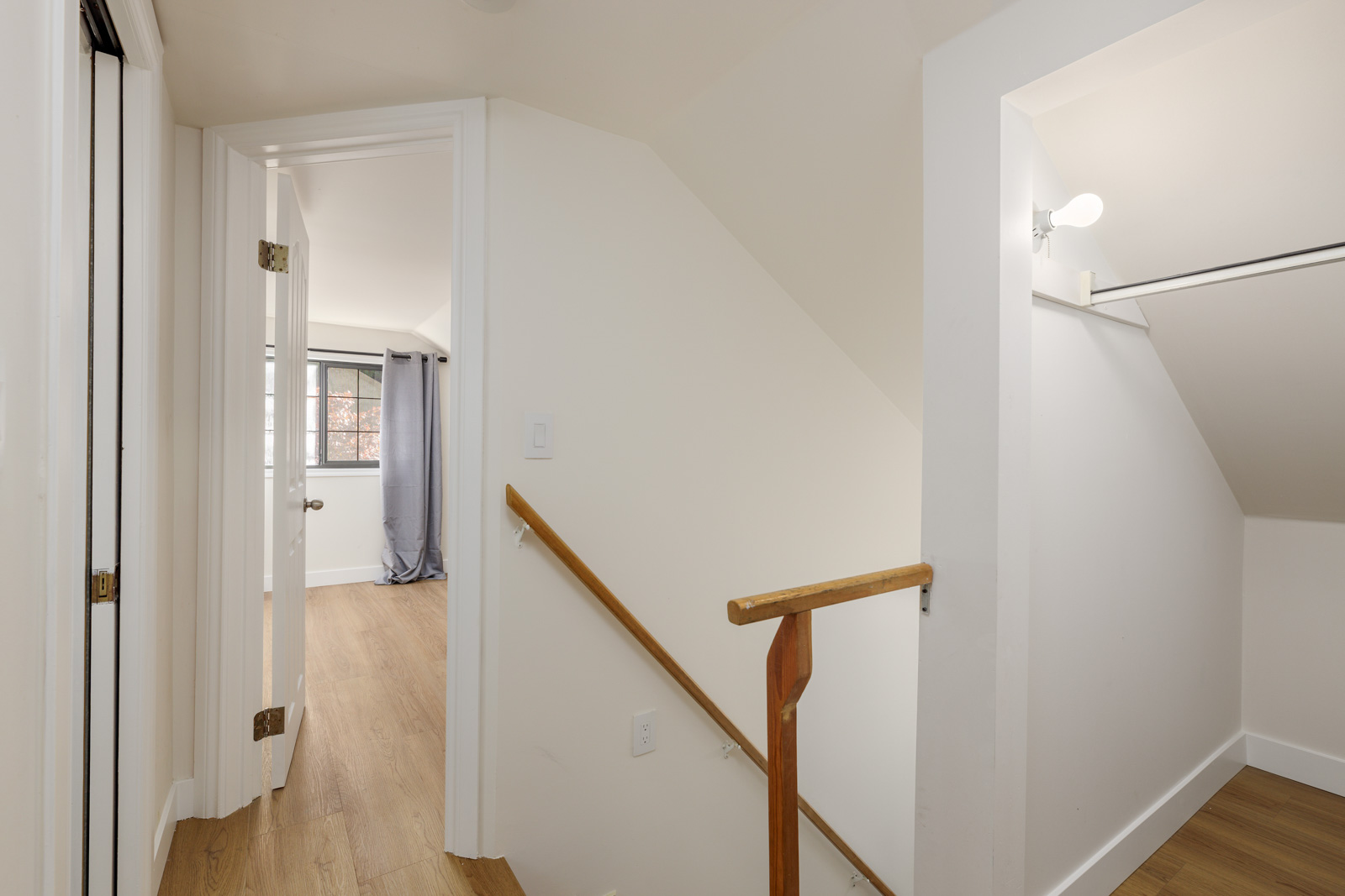 A hallway with wooden flooring leads to a room with a window and curtains. There's a stairway with a wooden handrail and a closet with an overhead light on the right.