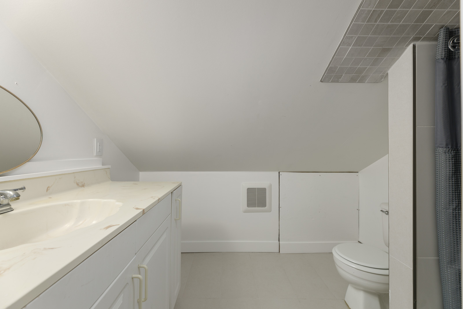 Bathroom with a sloped ceiling, a single sink on a marble countertop, and a toilet. There's a small vent on the wall and a shower curtain on the right.