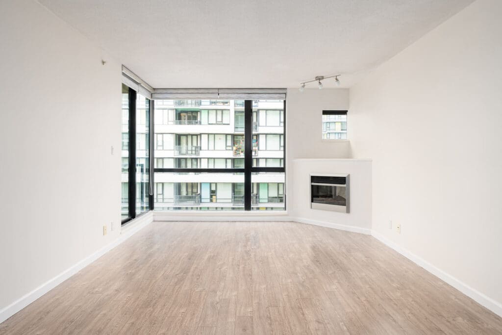 Empty modern apartment living room with light wood floors, large windows, and a corner fireplace.