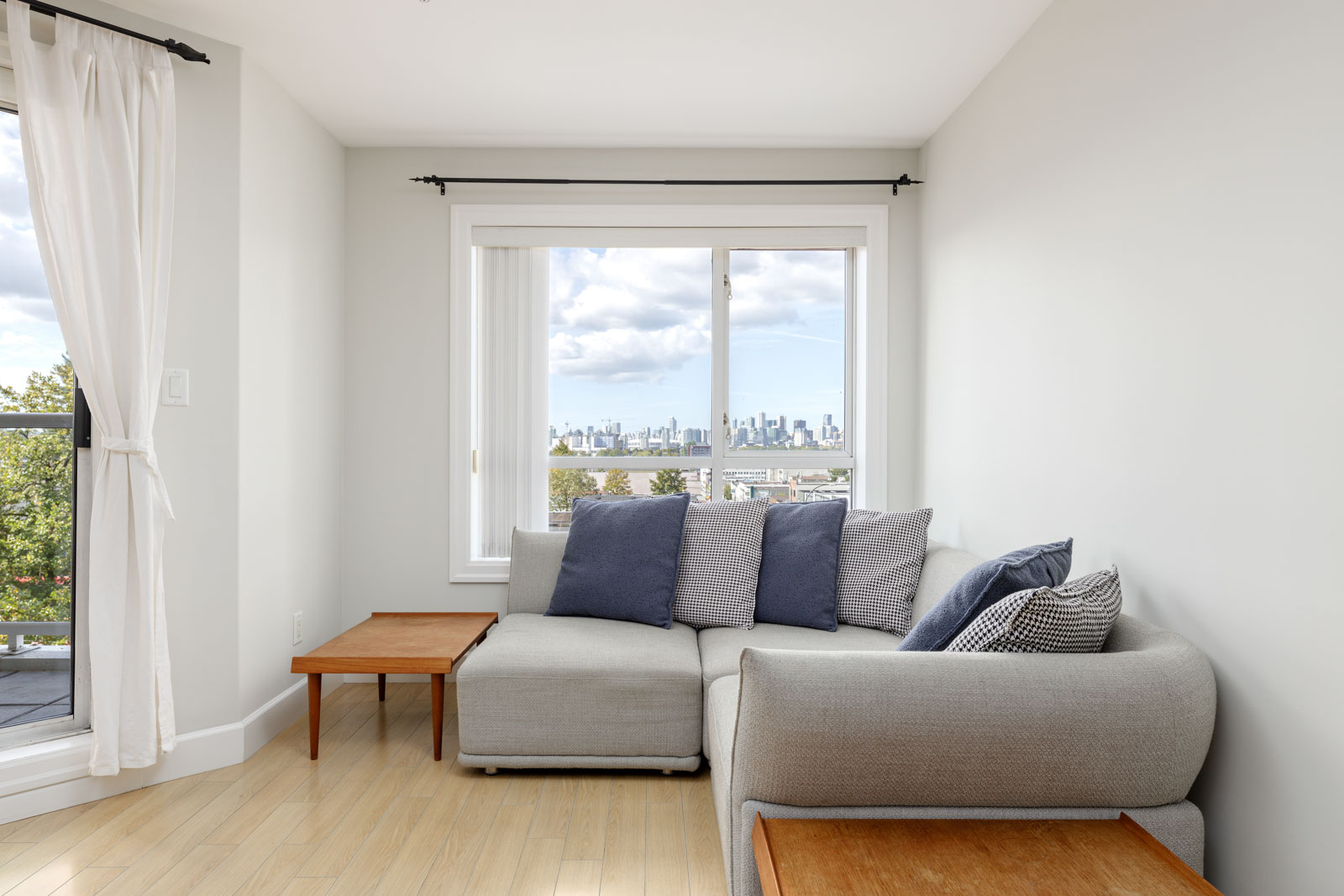 A cozy living room with a gray sectional sofa, wooden coffee tables, and a window view of a city skyline. White curtains frame a glass door to a balcony.