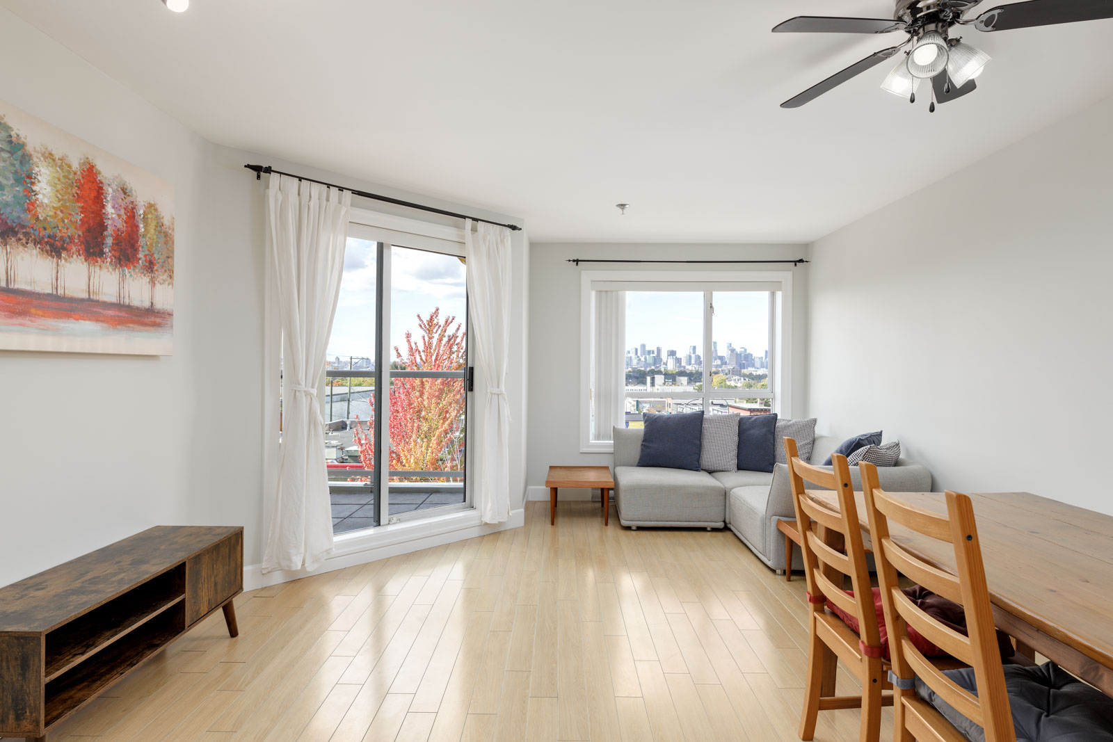 Bright living room with wooden floor, a gray sectional sofa, dining table with chairs, and large window overlooking a city skyline. A colorful landscape painting hangs on the wall.