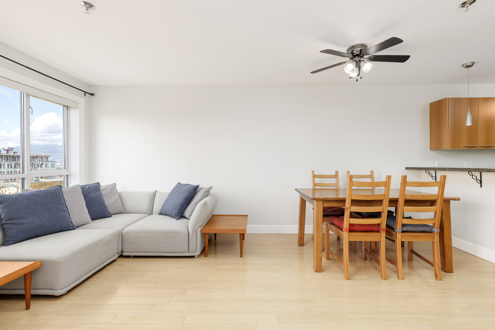 A bright living and dining area with a light gray sectional sofa, wooden coffee table, and a wooden dining set. A ceiling fan is above, and a large window provides natural light.