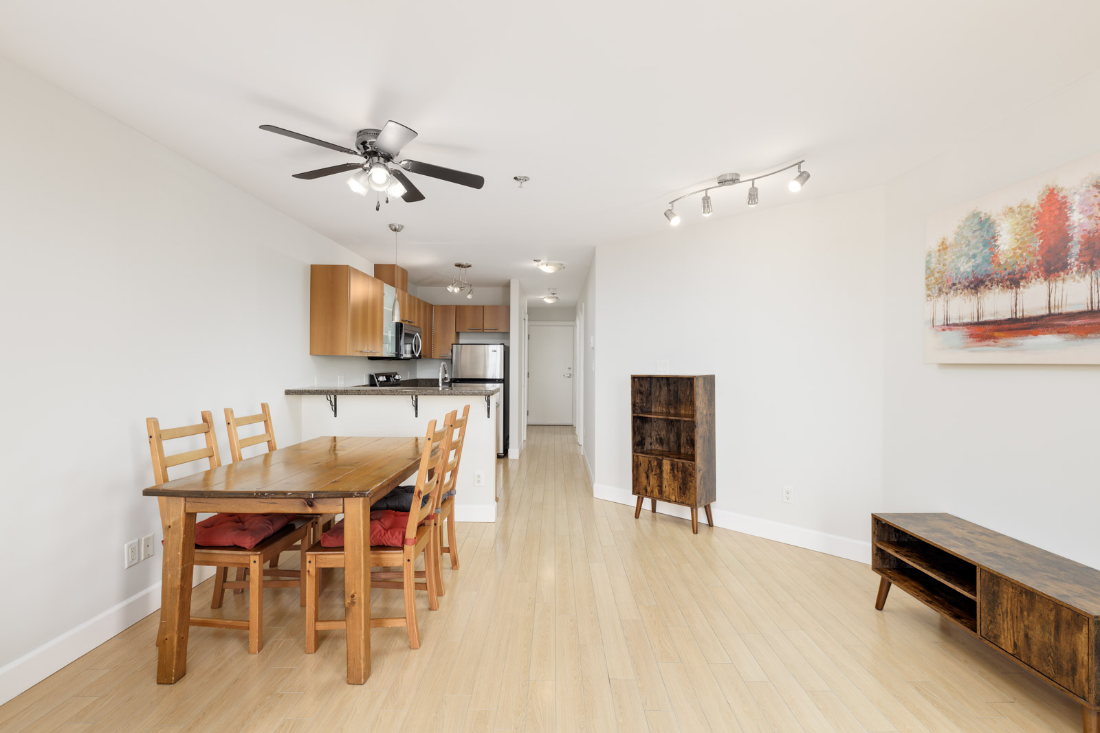 Simple dining and kitchen area with wooden table, chairs, and cabinets. Ceiling fans, light fixtures, and colorful tree artwork adorn the space.