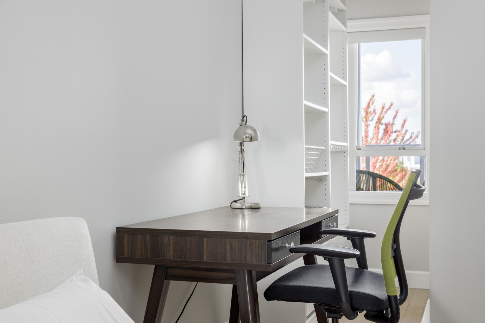 Minimalist home office with a wooden desk, modern lamp, and black swivel chair. White shelving and a window with a partial view of trees in the background.