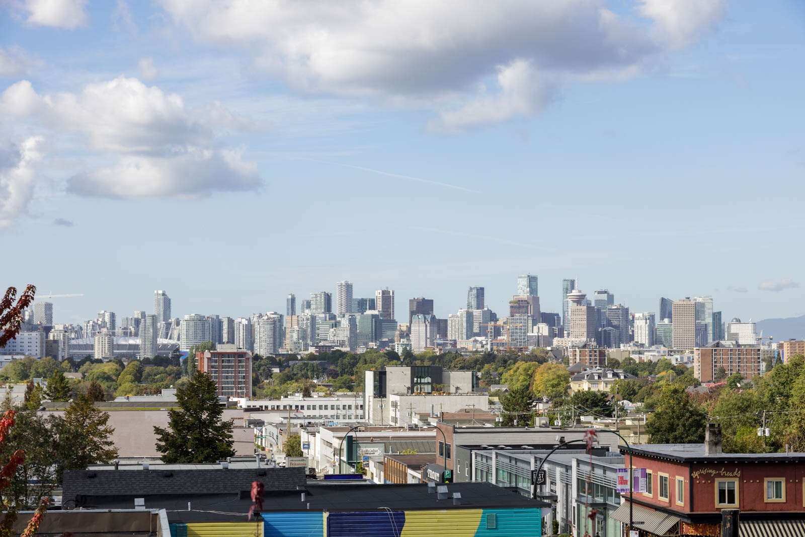 Skyline of a city with modern high-rise buildings under a partly cloudy sky; in the foreground, a colorful mural on a building and green trees are visible.