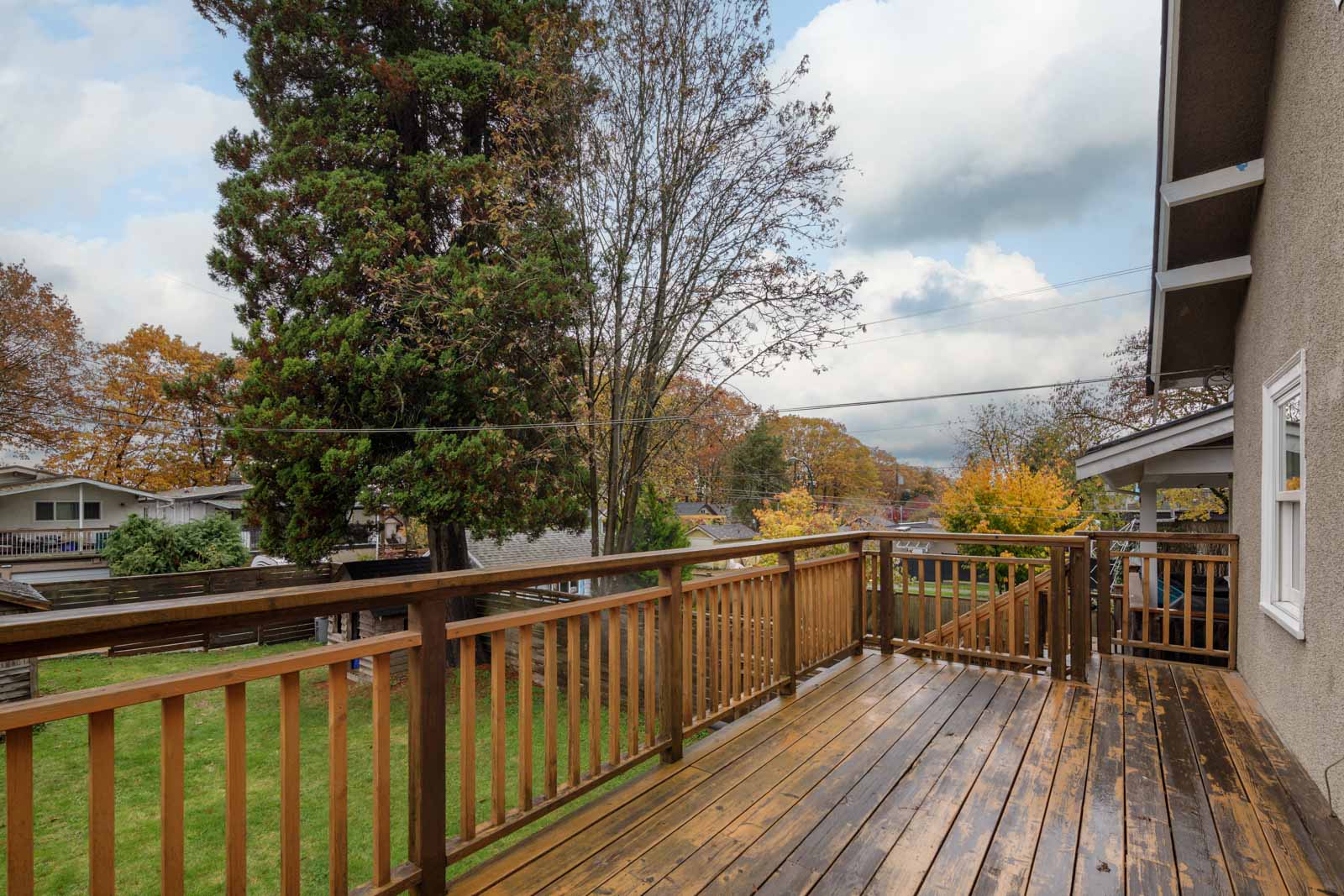 A wooden deck overlooks a green lawn with trees and neighboring houses under a cloudy sky.