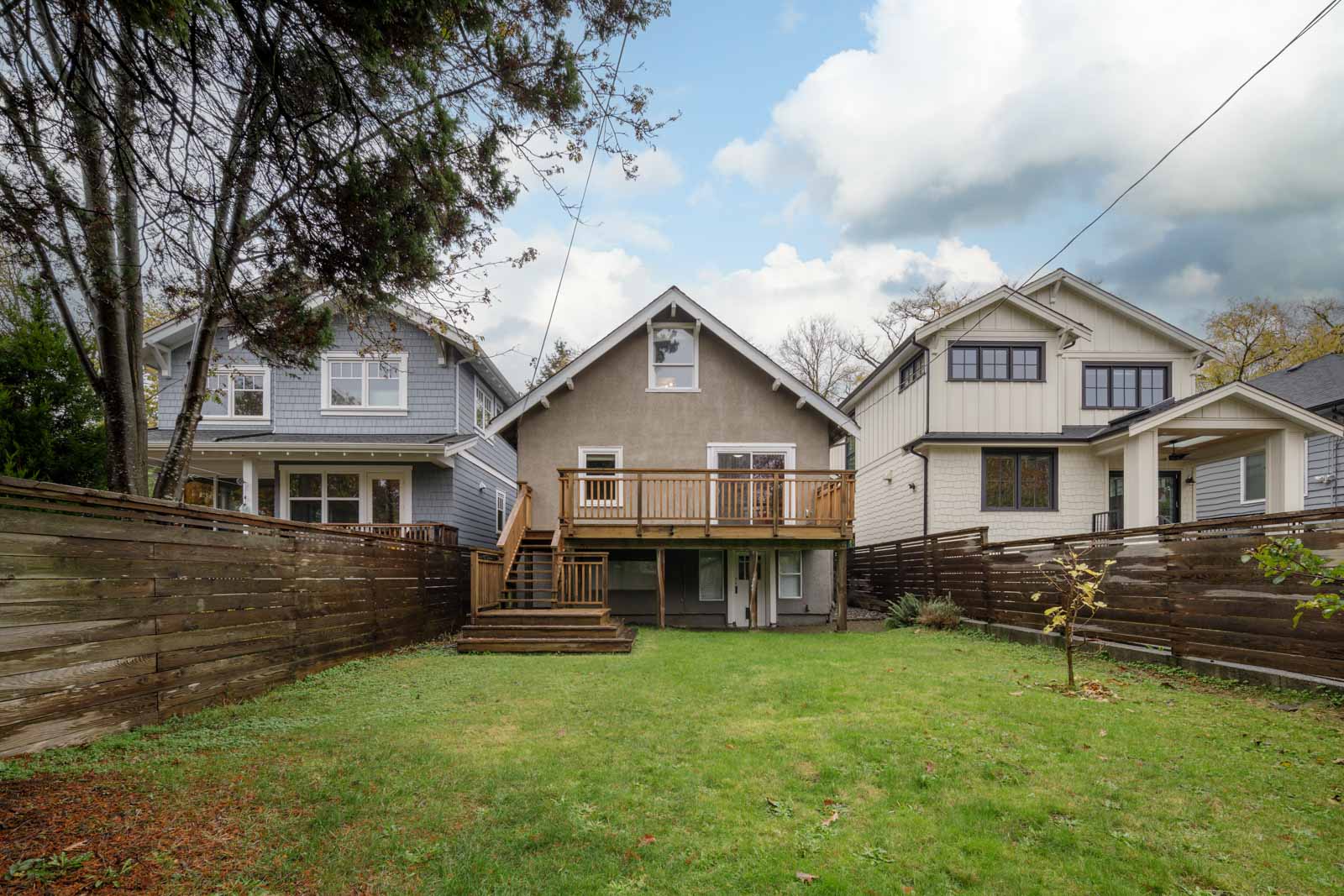 A view of three houses seen from the backyard, each with a wooden deck. The yard is grass-covered and bordered by wooden fences. The sky is partly cloudy.
