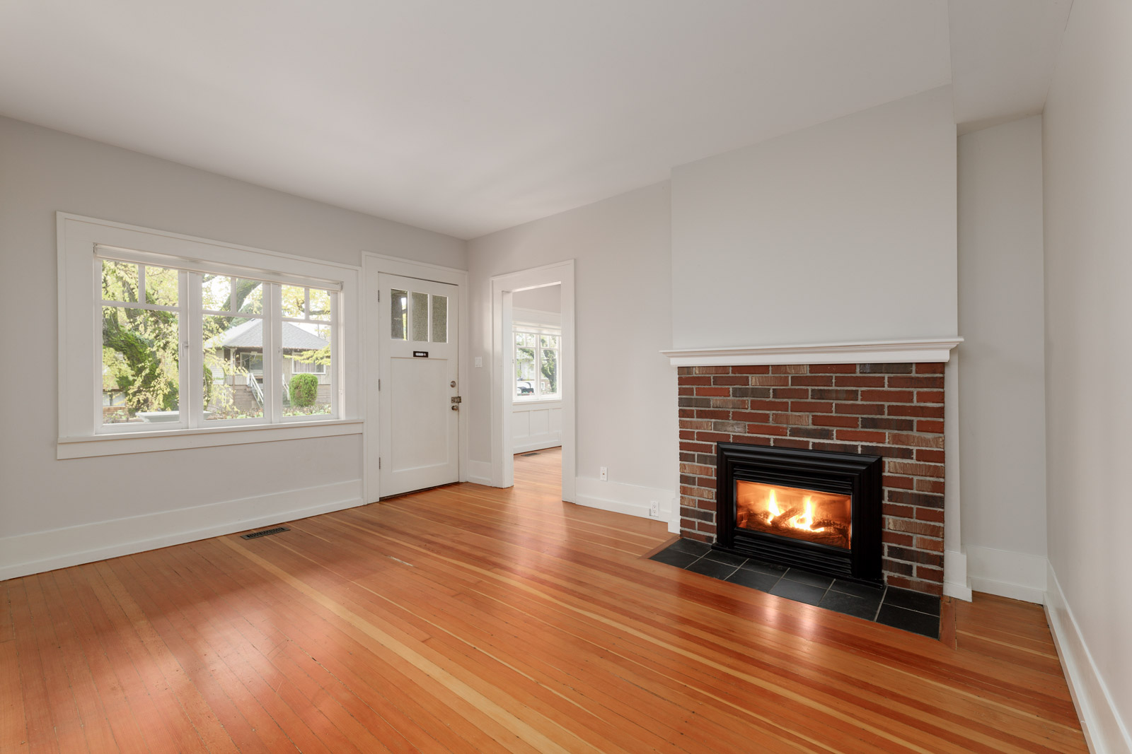 An empty living room with hardwood floors, a brick fireplace, a large window, and a white front door.
