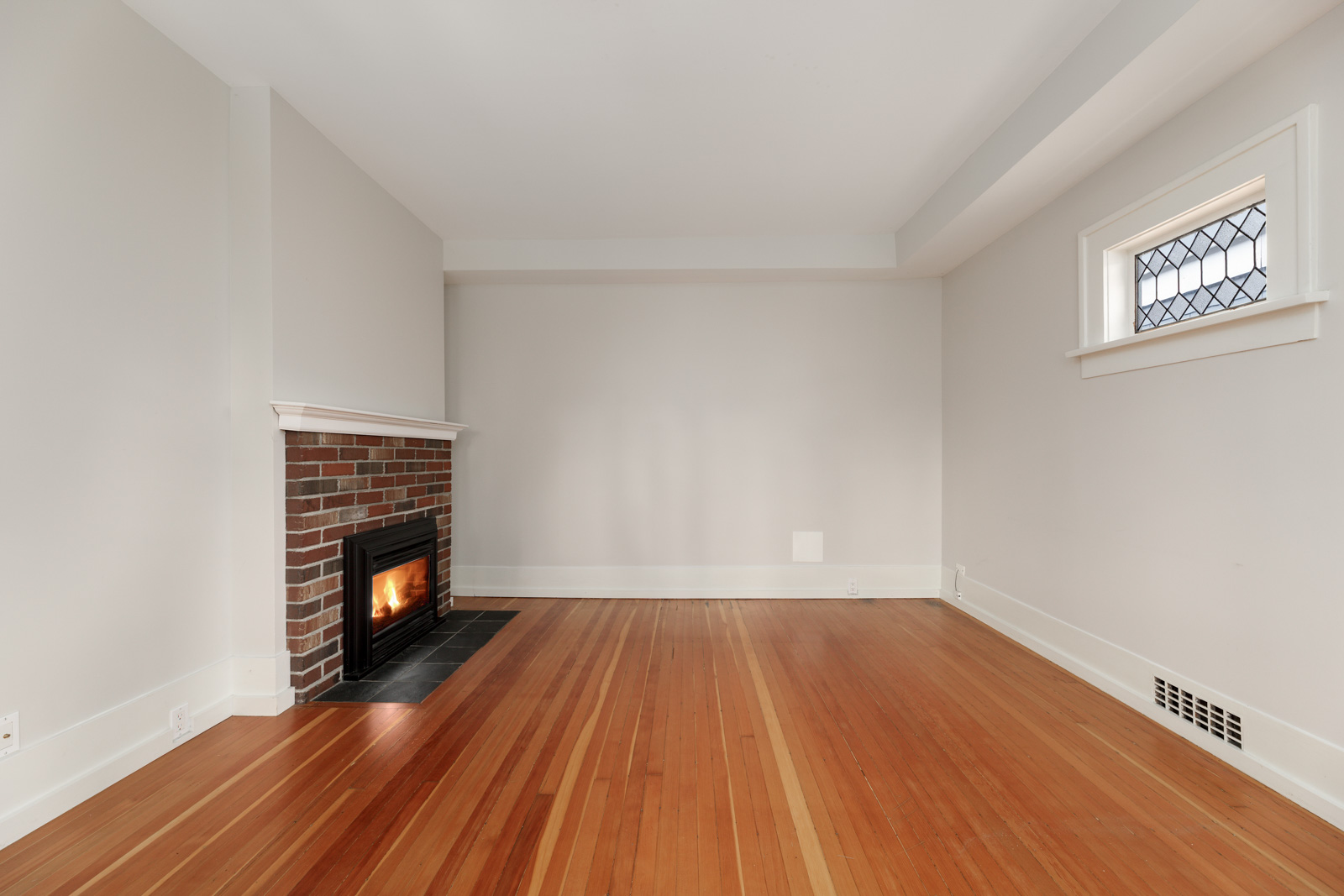 Empty room with hardwood flooring, a brick fireplace on the left, and a small window on the right.