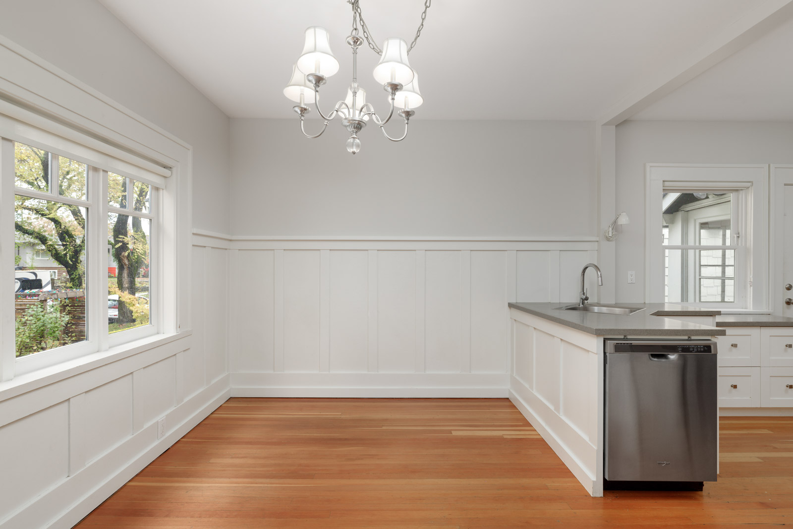 A bright kitchen with white cabinets, a stainless steel dishwasher, a sink, and a chandelier. Light wood flooring and a large window provide natural light.