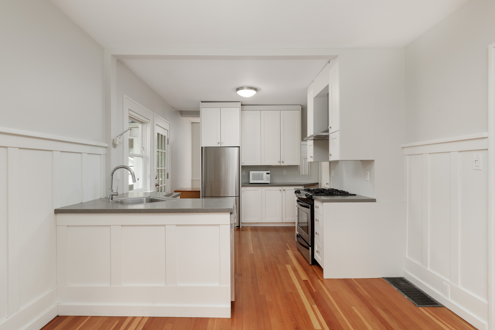 Modern kitchen with white cabinets, stainless steel appliances, and wooden flooring. The room features a center island with a sink and bright natural lighting.