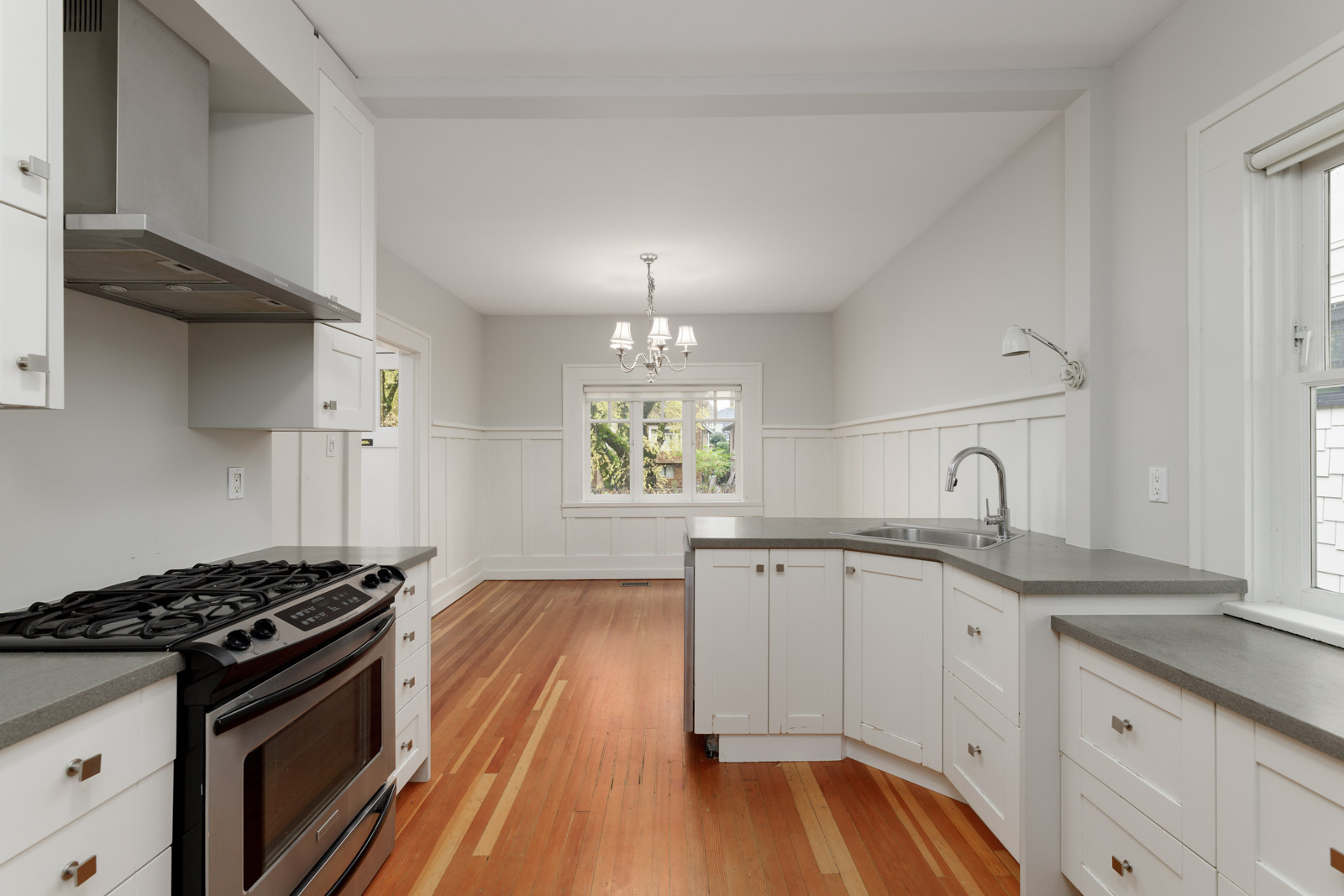 A modern kitchen with white cabinets and a gray countertop, featuring a stainless steel oven and a sink. Wooden flooring extends to a dining area with a window and chandelier.