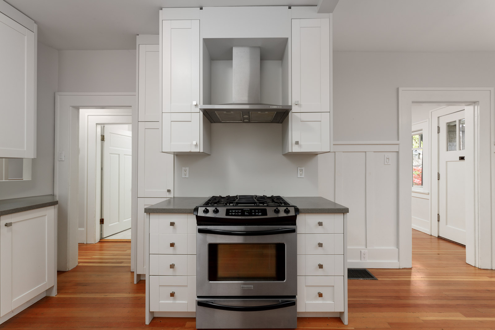 A modern kitchen with a stainless steel gas oven, white cabinets, and wooden flooring. There are doors on either side of the oven area.