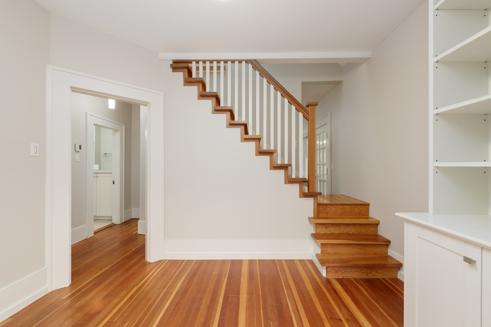 Interior view of a room with polished wood floors, a wooden staircase with white railings, built-in shelves on the right, and a doorway leading to another room.