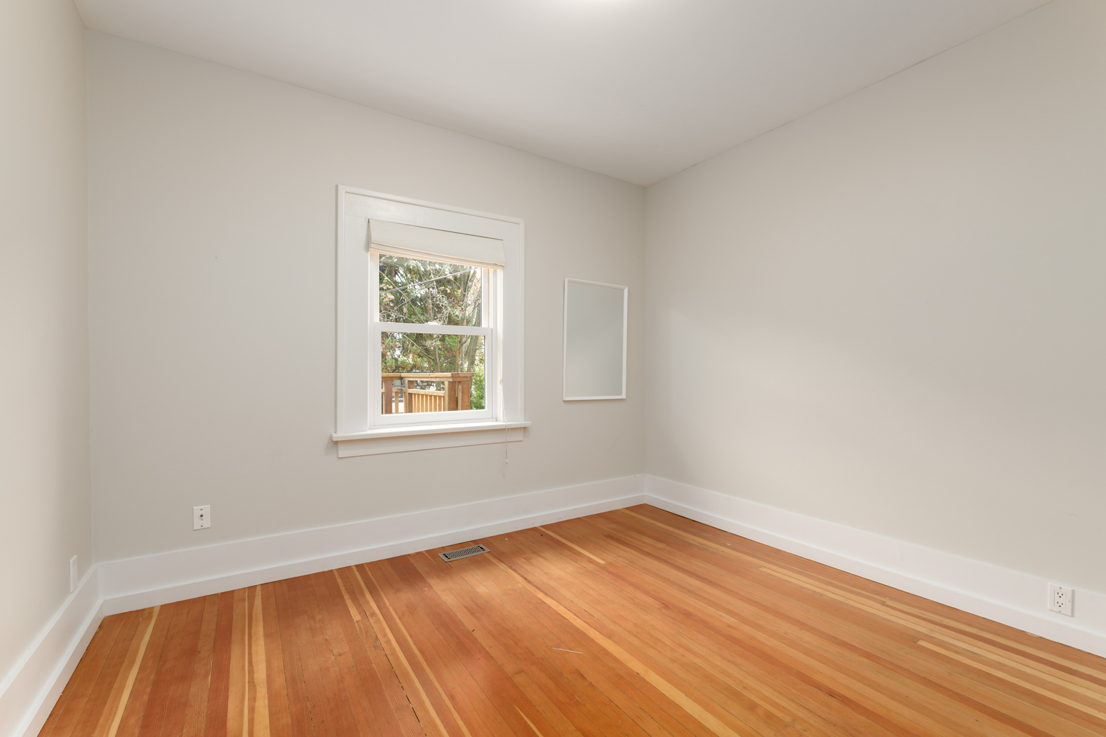 An empty room with light beige walls, a window with a view of trees, and wooden flooring. A rectangular mirror is on one of the walls.