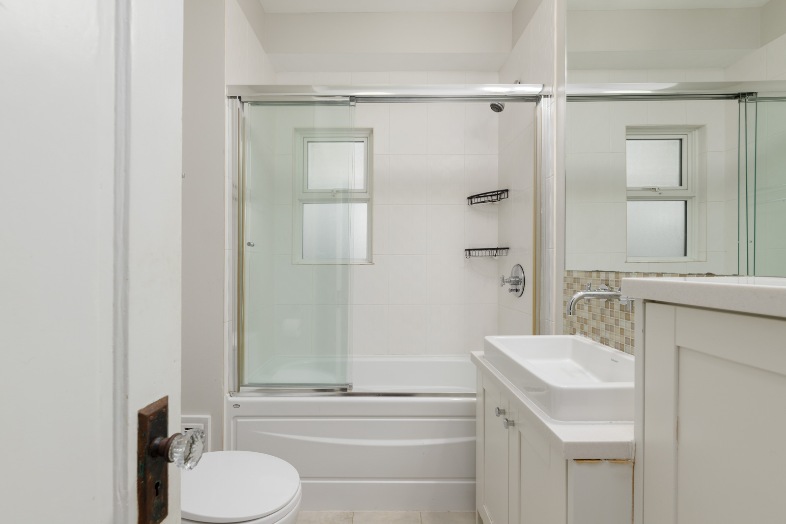 A clean, modern bathroom with a white bathtub and shower, glass doors, white sink with a cabinet, and a toilet. Two small windows provide natural light.