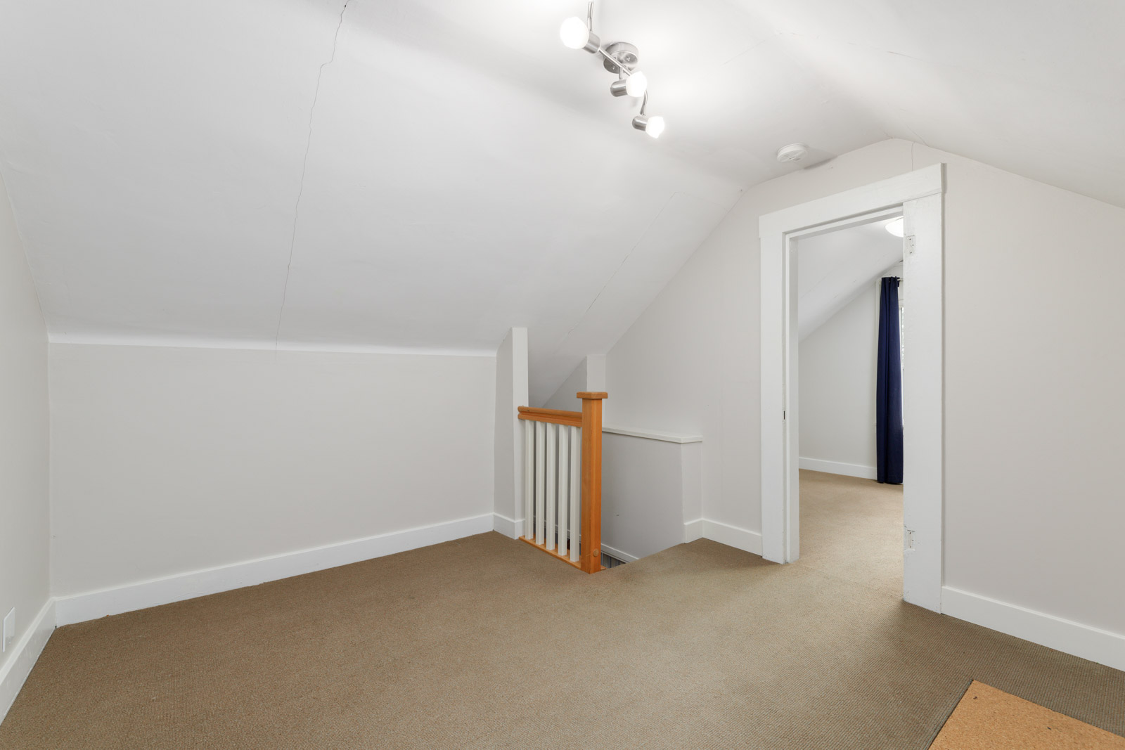 An empty, carpeted attic room with white walls and a ceiling light. A wooden staircase railing is visible, leading to an adjacent room with a dark curtain.