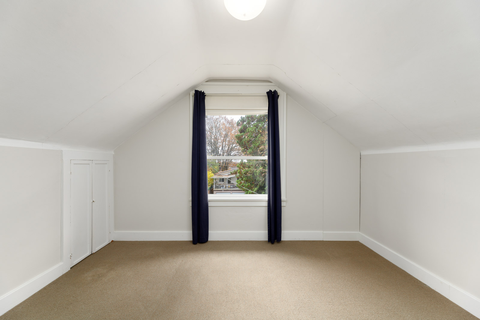 An empty room with white walls and a sloped ceiling, featuring a window with dark curtains, beige carpet, and a ceiling light.