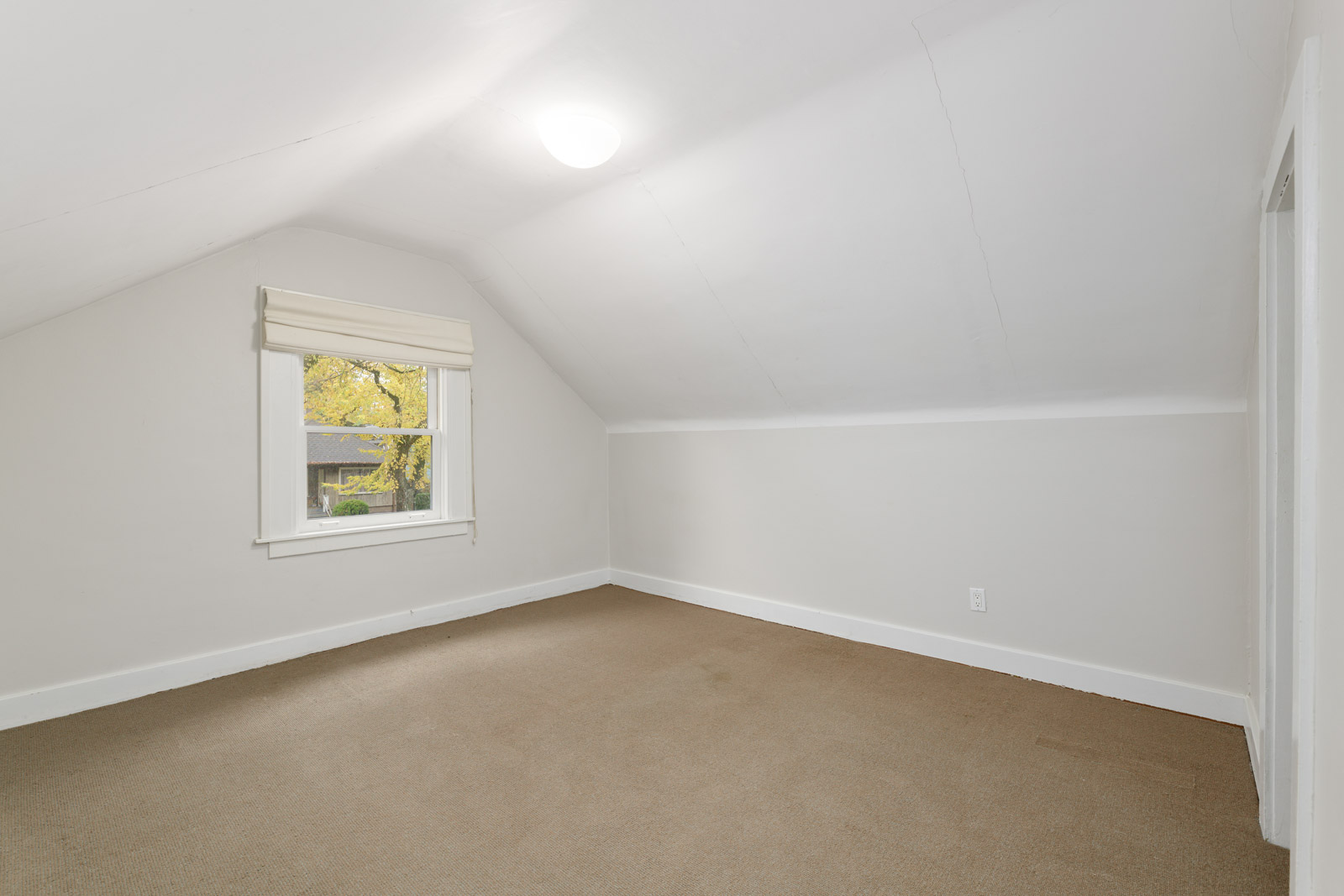 An empty room with beige carpet, white walls, and a sloped ceiling. A small window with a blind shows trees outside.