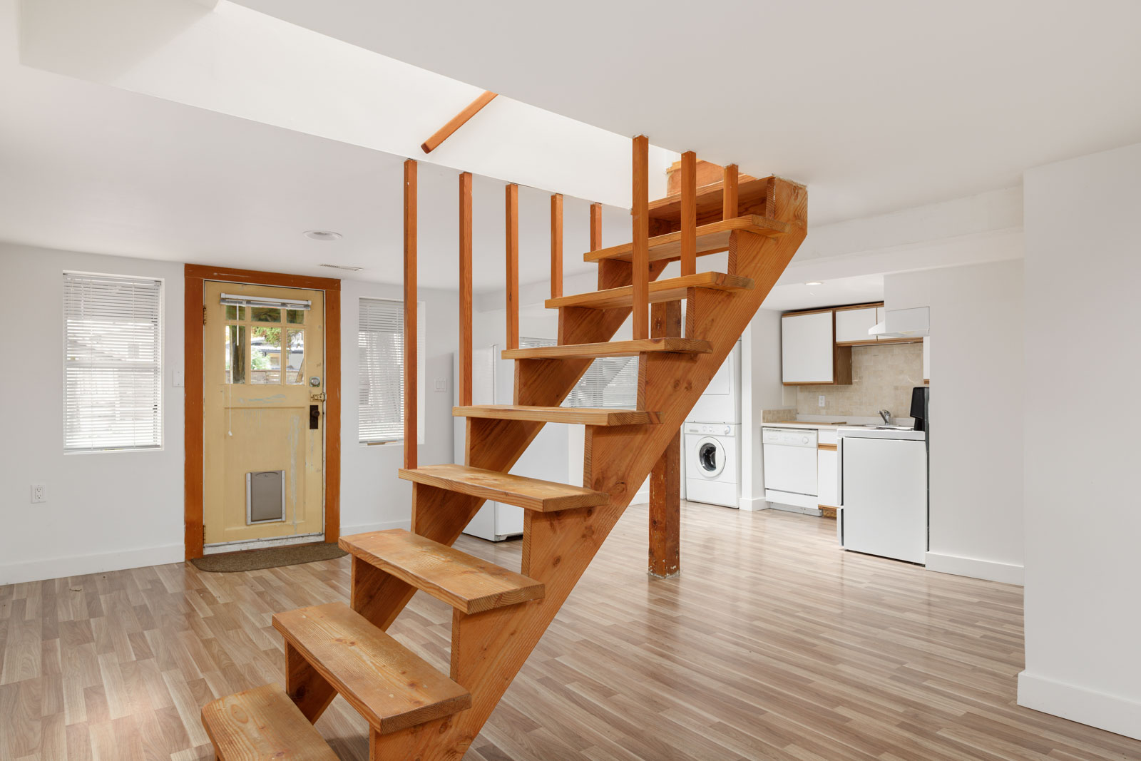 Interior view of a room with wooden stairs leading to an upper level, light wood flooring, a yellow door with a pet flap, and a kitchen area with appliances in the background.