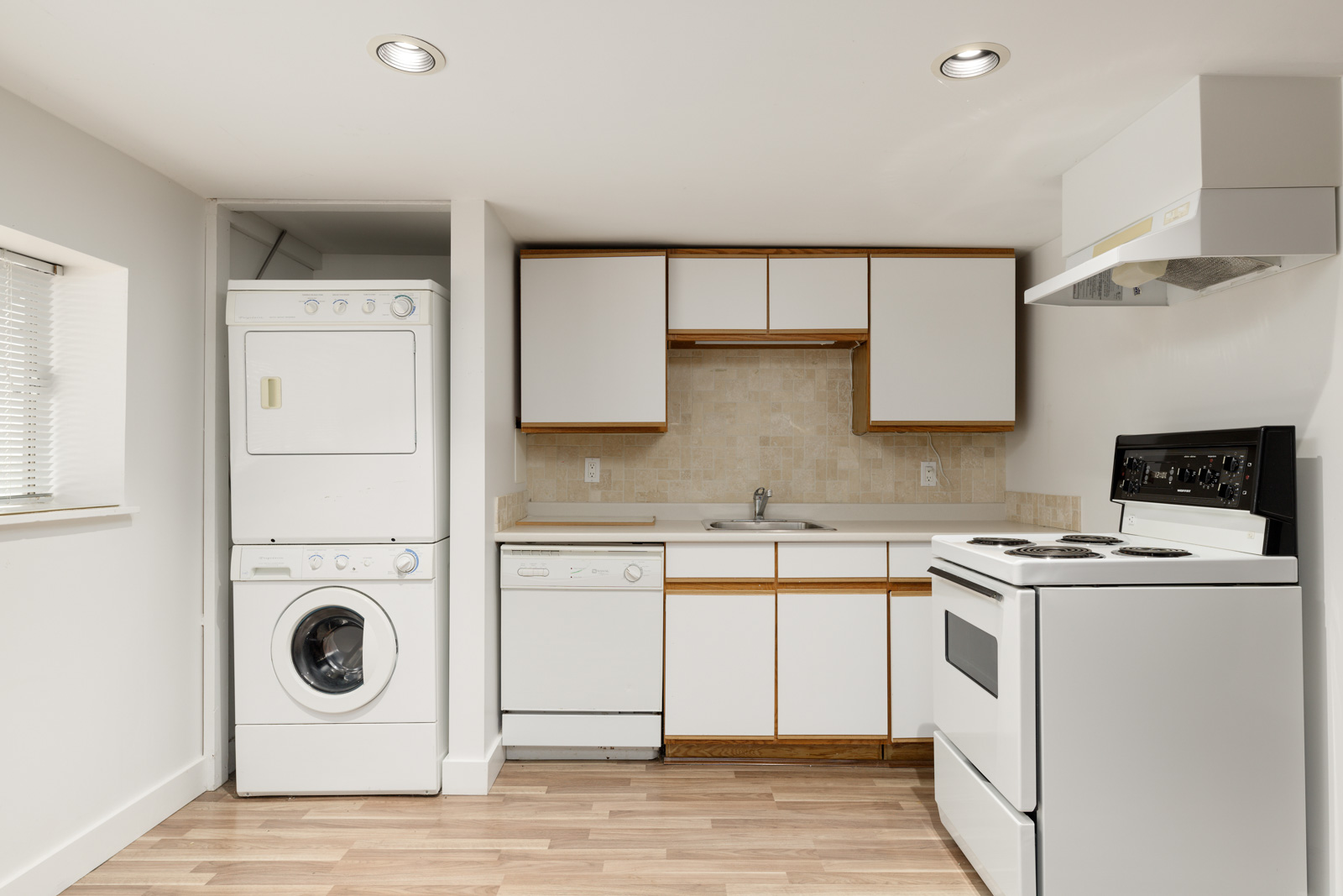 Compact kitchen with white cabinets, sink, dishwasher, stove, and a stacked washer and dryer set against a light wood floor.