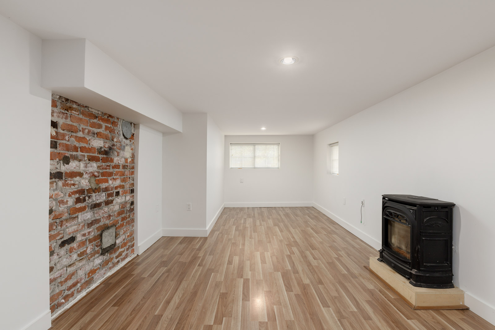 A basement room with faux wood flooring, exposed brick wall, wood stove, and recessed lighting. A small window is centered on the back wall.