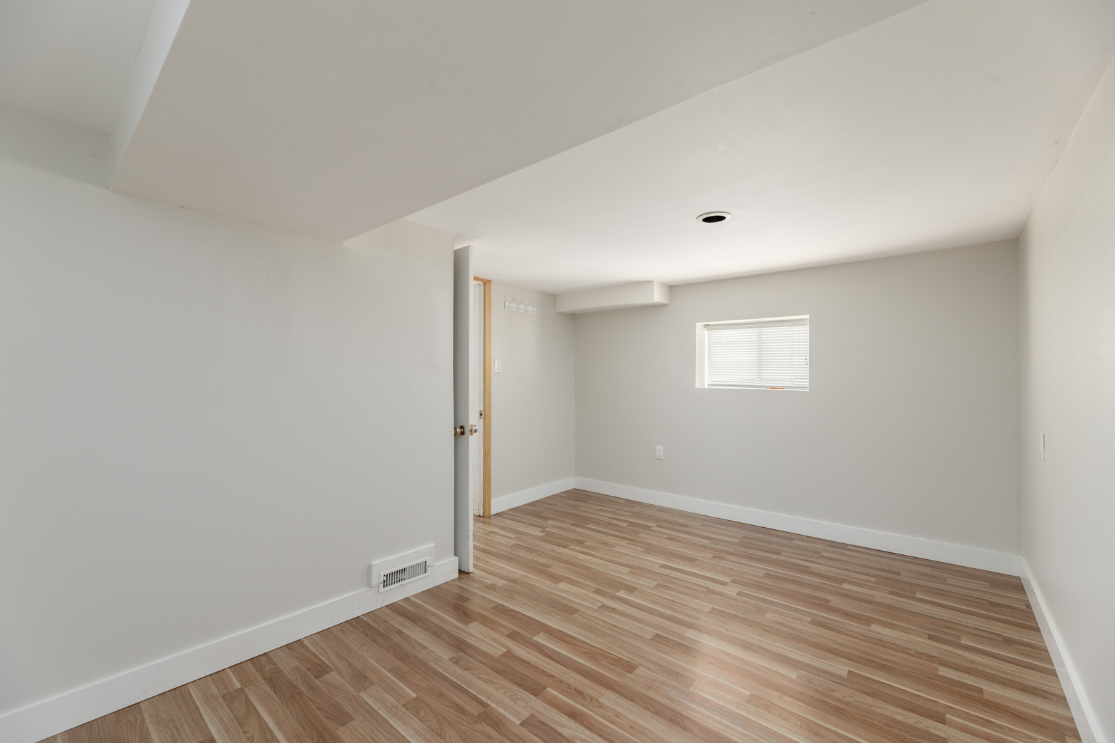 Empty room with light wood flooring, white walls, and a small window. A door is partially open. There's a ceiling vent and recessed lighting.