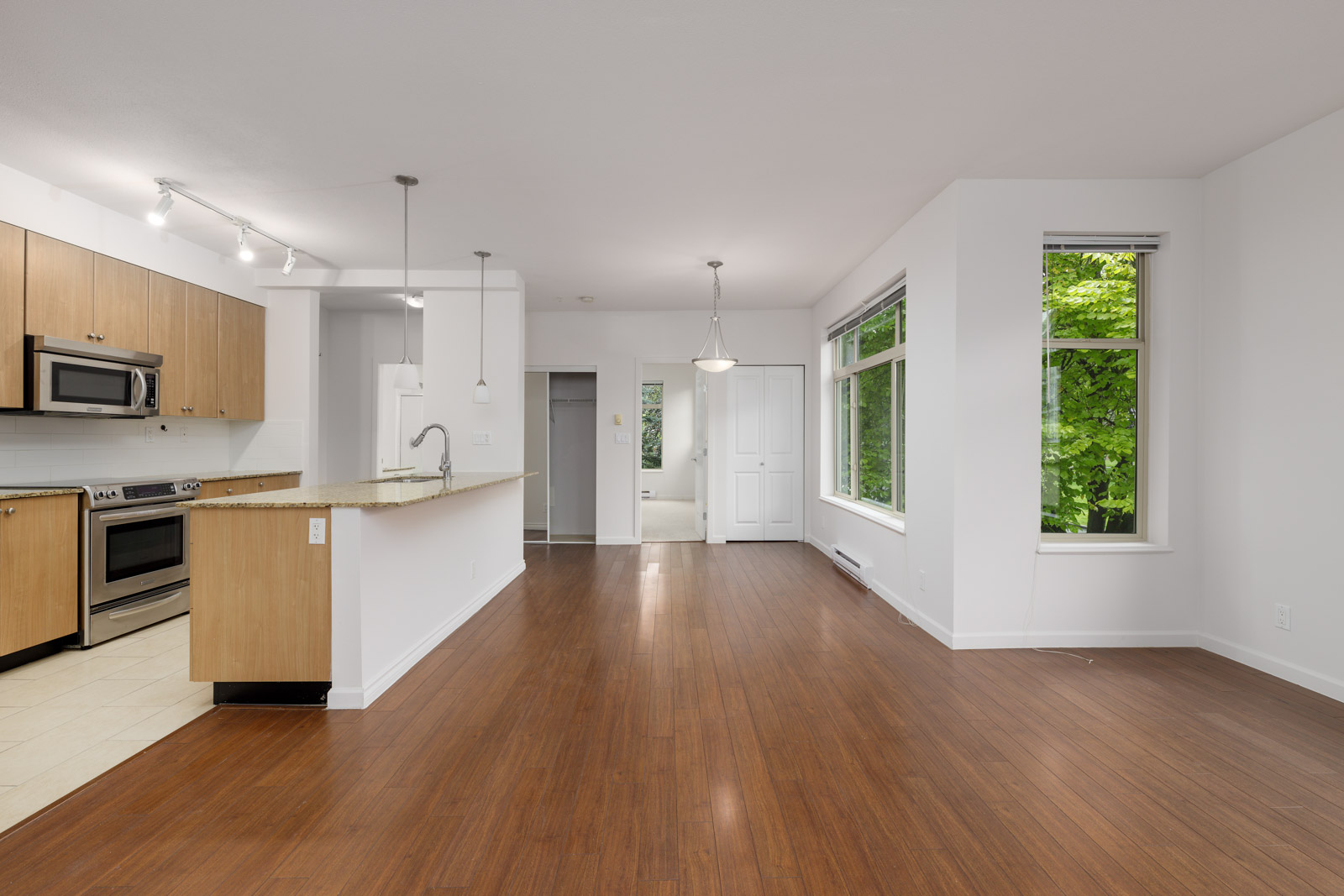Modern open-plan kitchen and living area with wooden floors, stainless steel appliances, and large windows offering a view of greenery.
