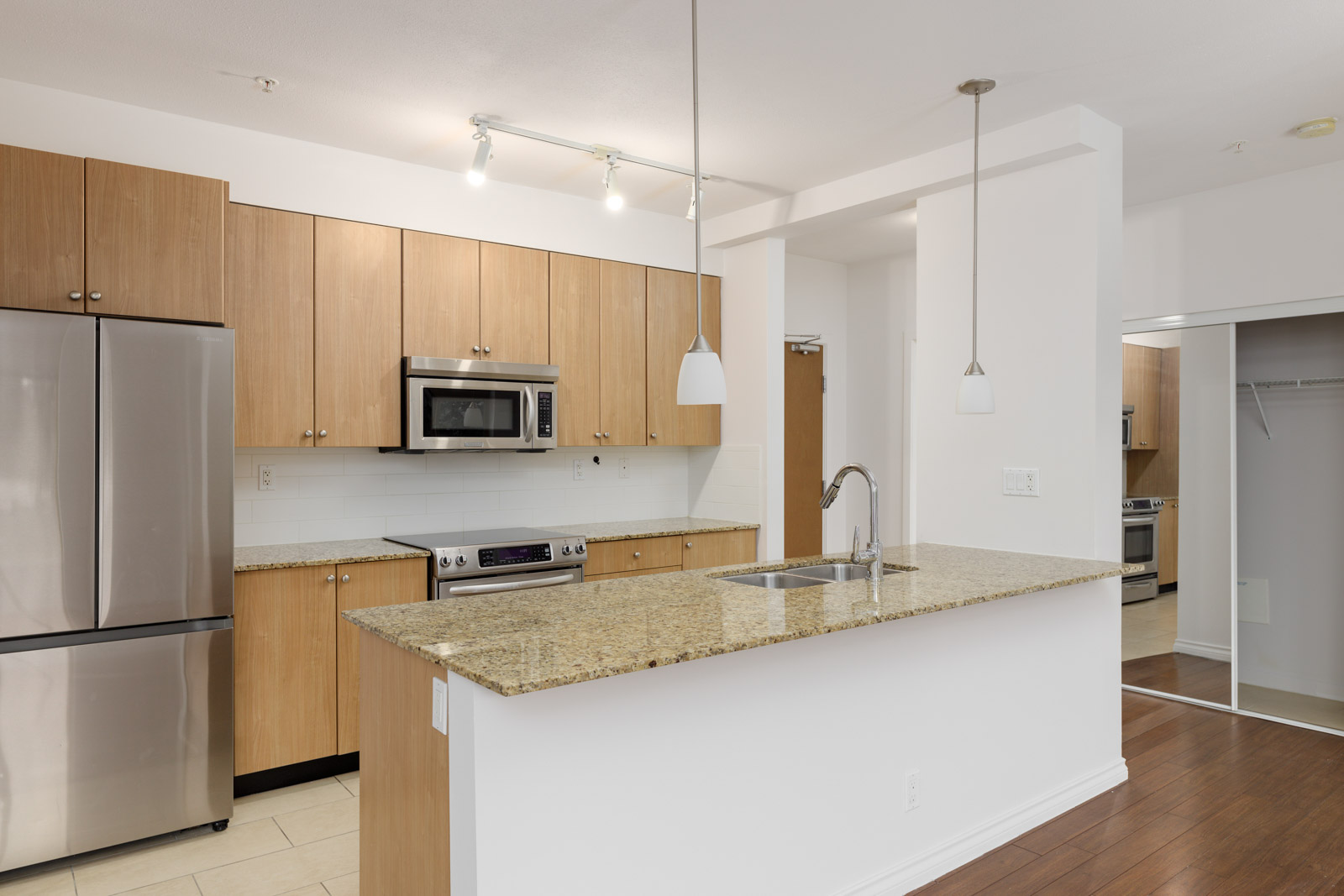 Modern kitchen with stainless steel appliances, light wooden cabinets, a granite countertop island, and hardwood floors. Track lighting and pendant lights are installed above the island.