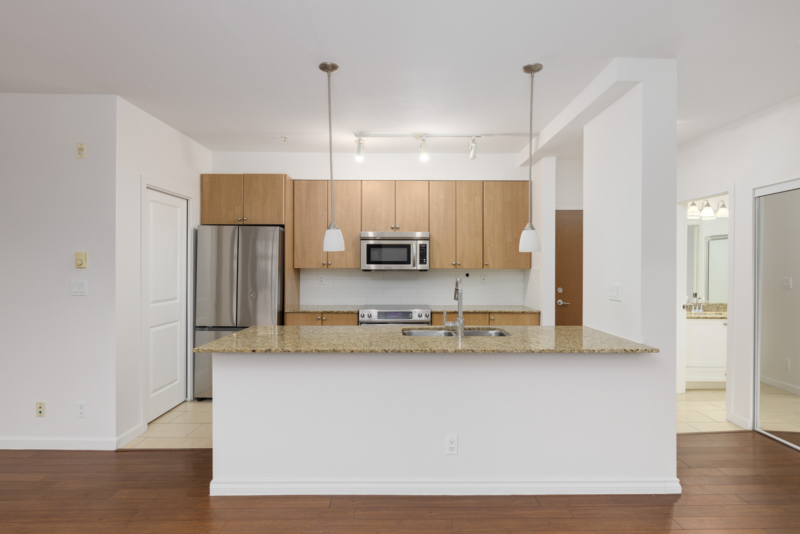 Modern kitchen with wooden cabinets, a granite countertop island, a stainless steel refrigerator, and a built-in microwave. Two pendant lights hang above the island.