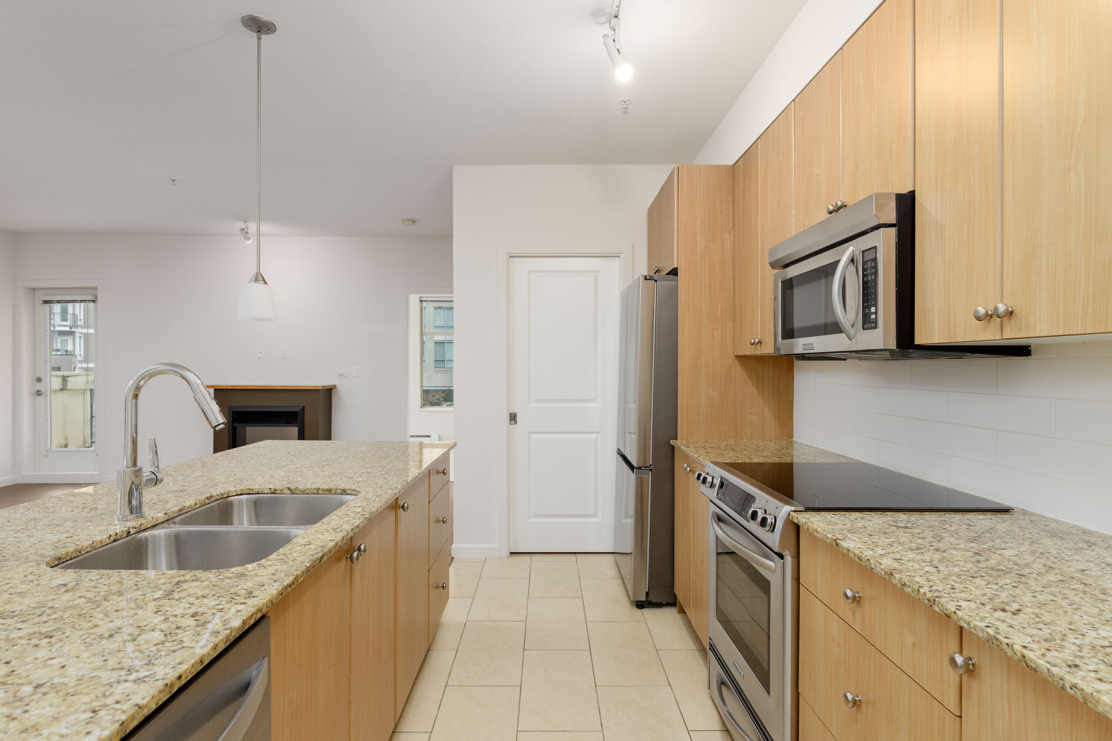Modern kitchen with granite countertops, stainless steel appliances, a double sink, and light wood cabinets. Cream-colored tile flooring and a view of the living area.