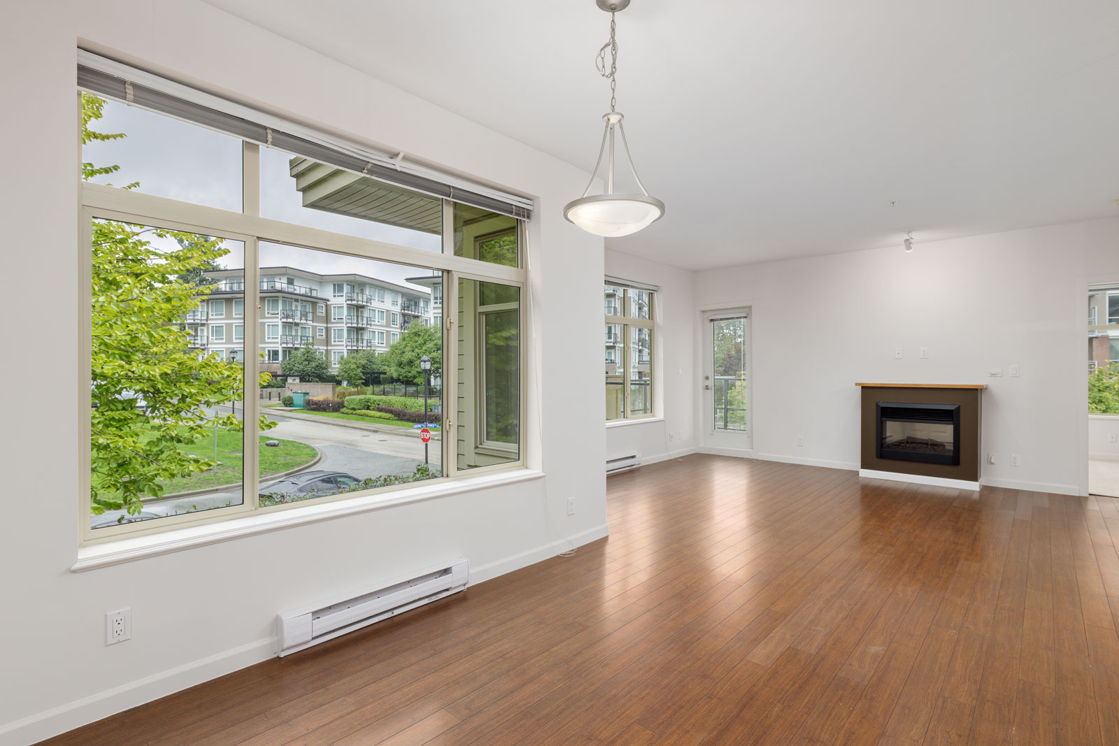Empty living room with large windows, wooden floors, a ceiling light, and a fireplace. View of apartment buildings and greenery outside.