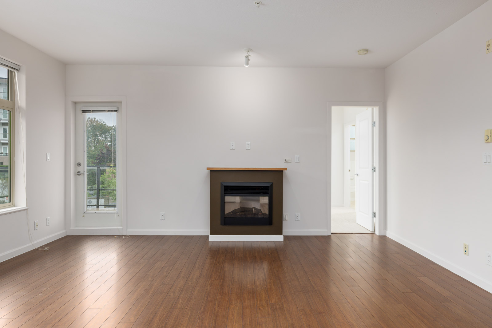 Empty room with wooden flooring, a central fireplace, and a door leading to a balcony. White walls and ceiling with two open doorways on the right.