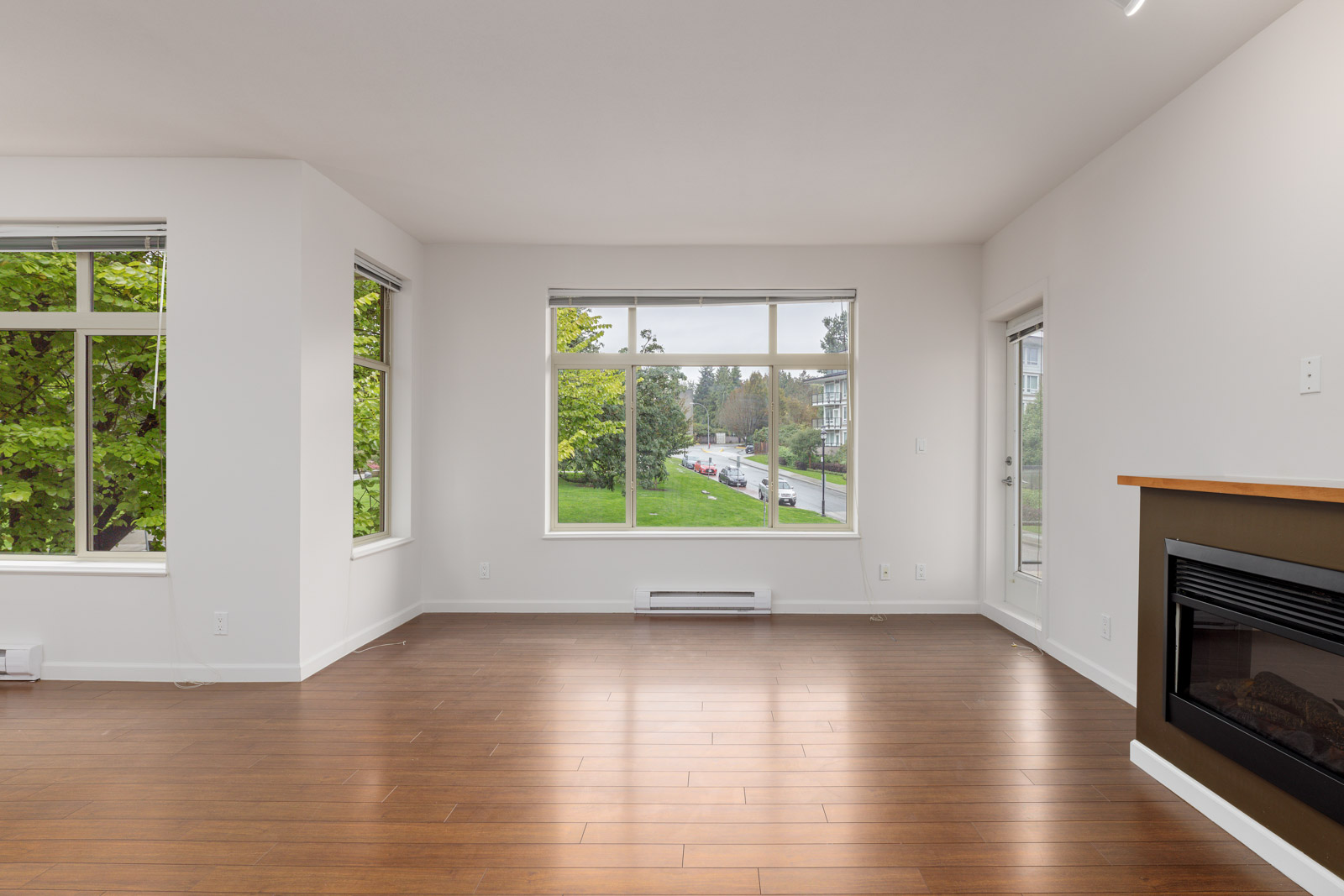 Spacious, unfurnished room with wooden floors, a large window overlooking a street, and a fireplace on the right.