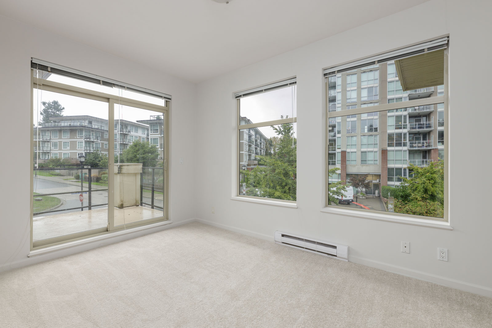 Empty room with large windows and beige carpet, overlooking an apartment complex and greenery outside.