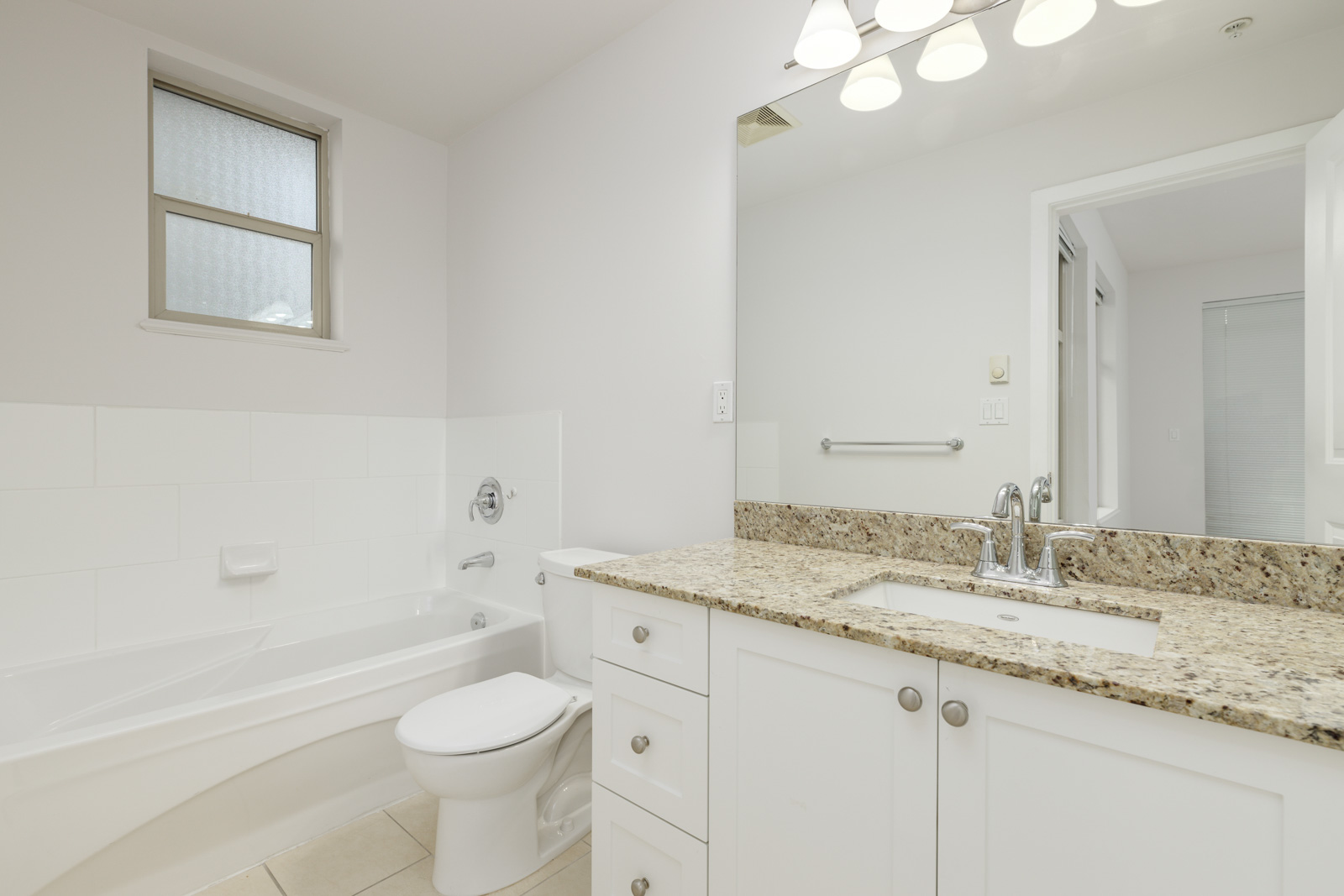 A clean white bathroom with a granite countertop, sink, bathtub, toilet, and a well-lit mirror above the sink. A small window is above the bathtub.