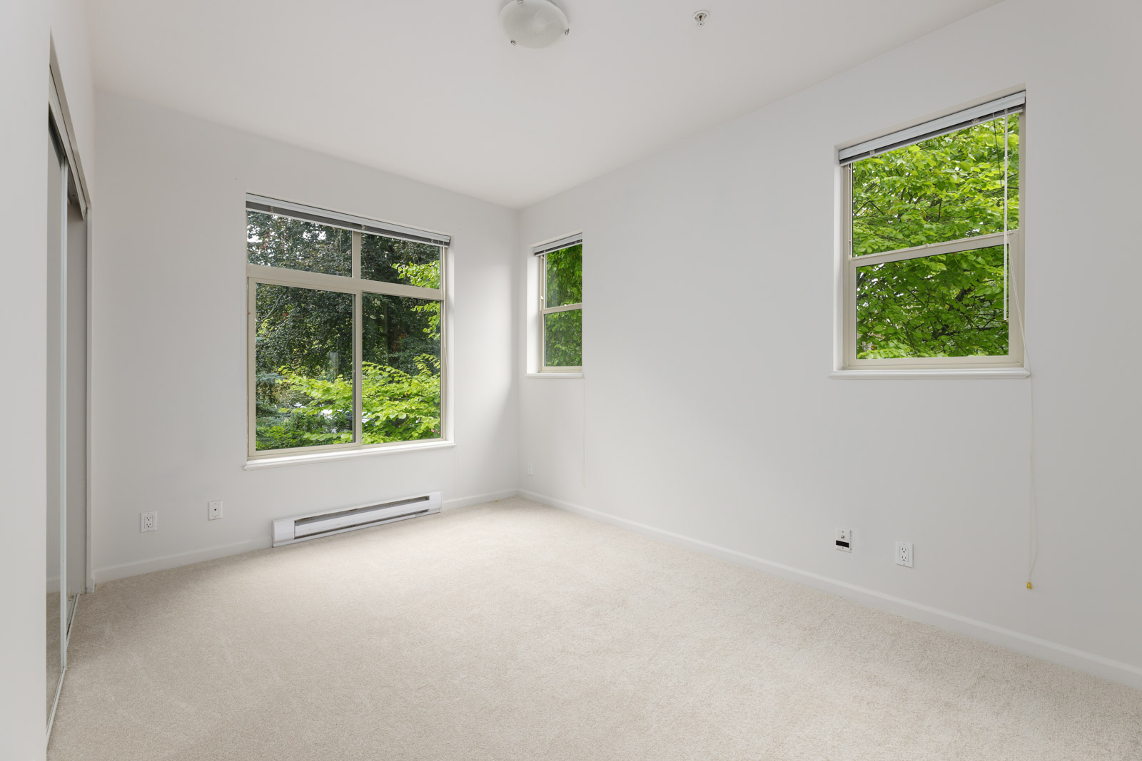 Empty room with light gray walls, beige carpet, and two windows showing green trees outside. Ceiling light fixture and baseboard heater are visible.