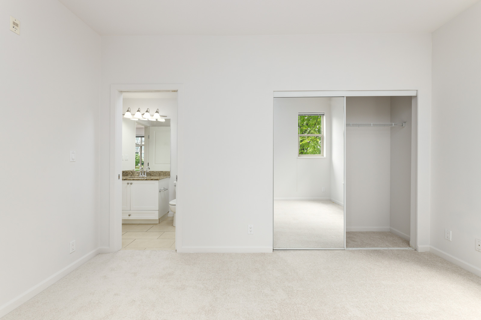 Empty room with beige carpet, white walls, and a mirrored closet door. Doorway on the left leads to a bathroom with a vanity and lights. Small window provides natural light.