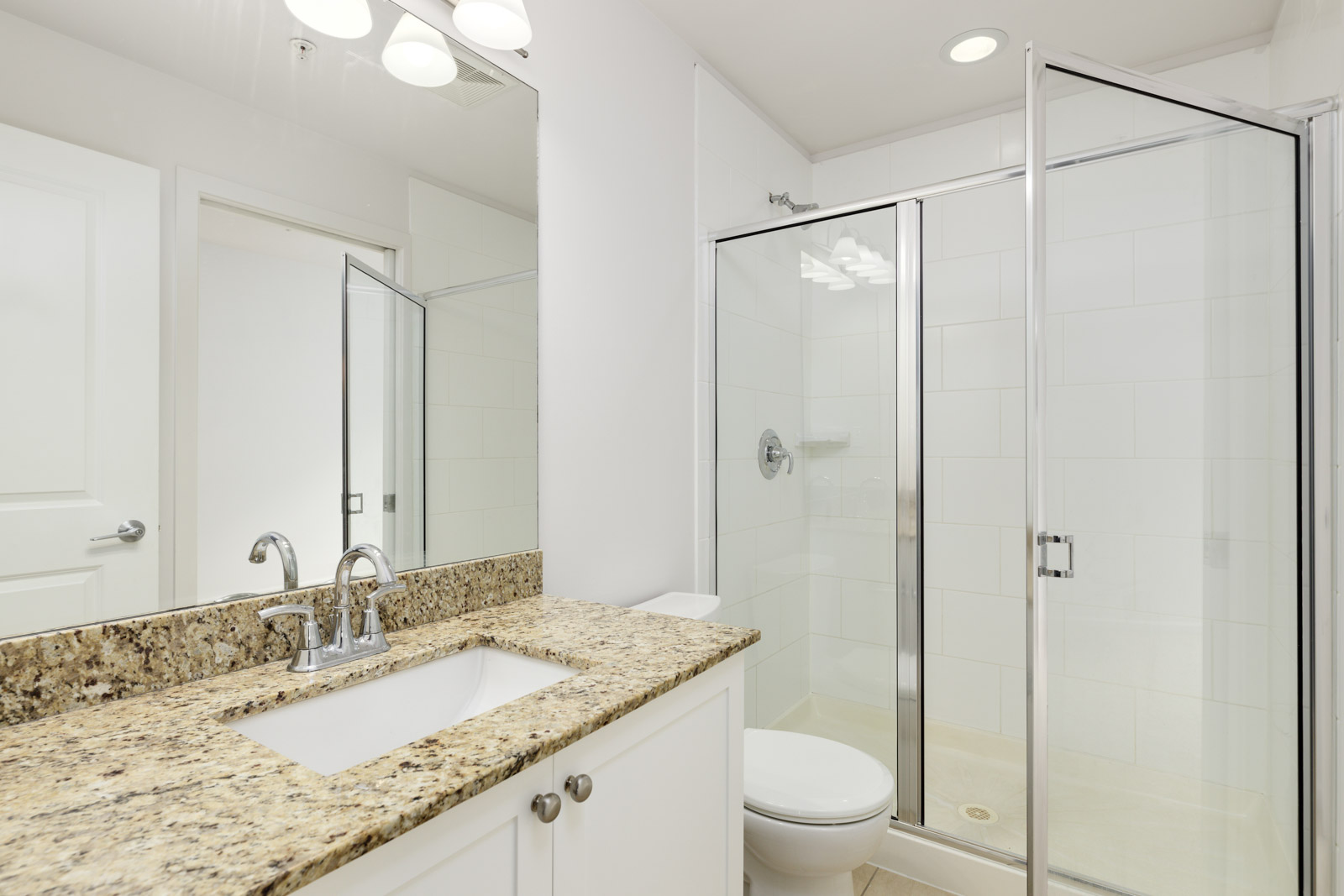 Bathroom with a glass-enclosed shower, granite countertop with a sink, and a large mirror. White cabinets are below the sink.
