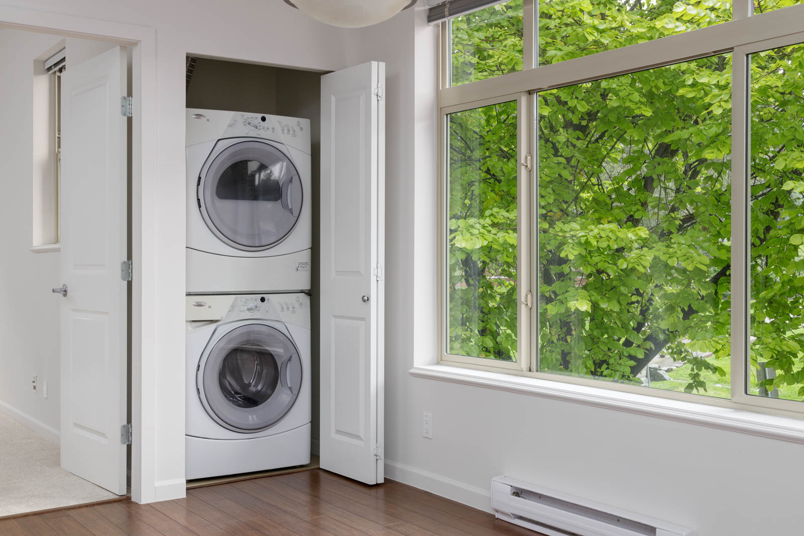 A stacked washer and dryer are in a closet next to a large window with a view of green trees.