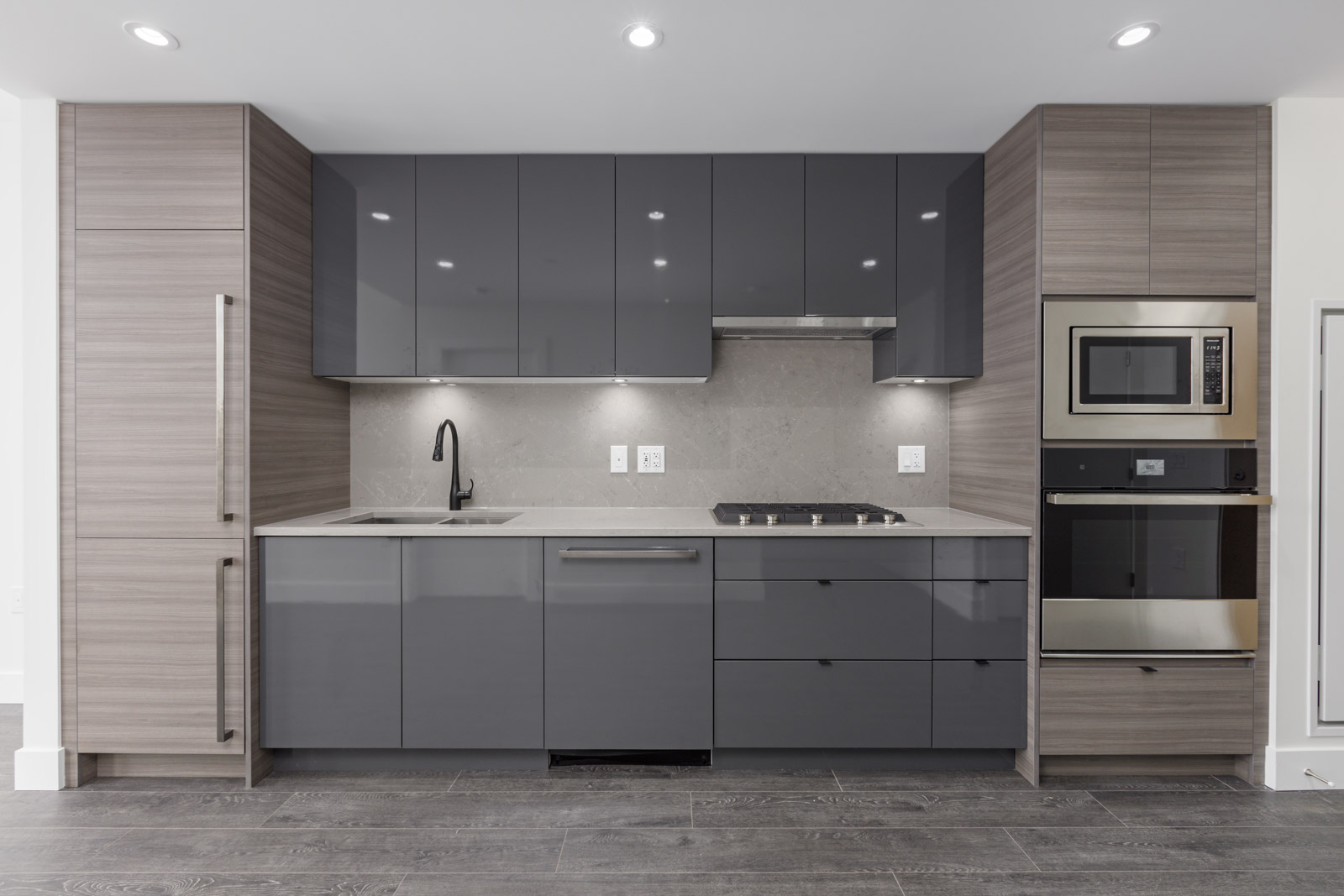 Modern kitchen with dark gray cabinets, built-in oven, microwave, and sleek appliances. Quartz countertop and light wood flooring complete the look.