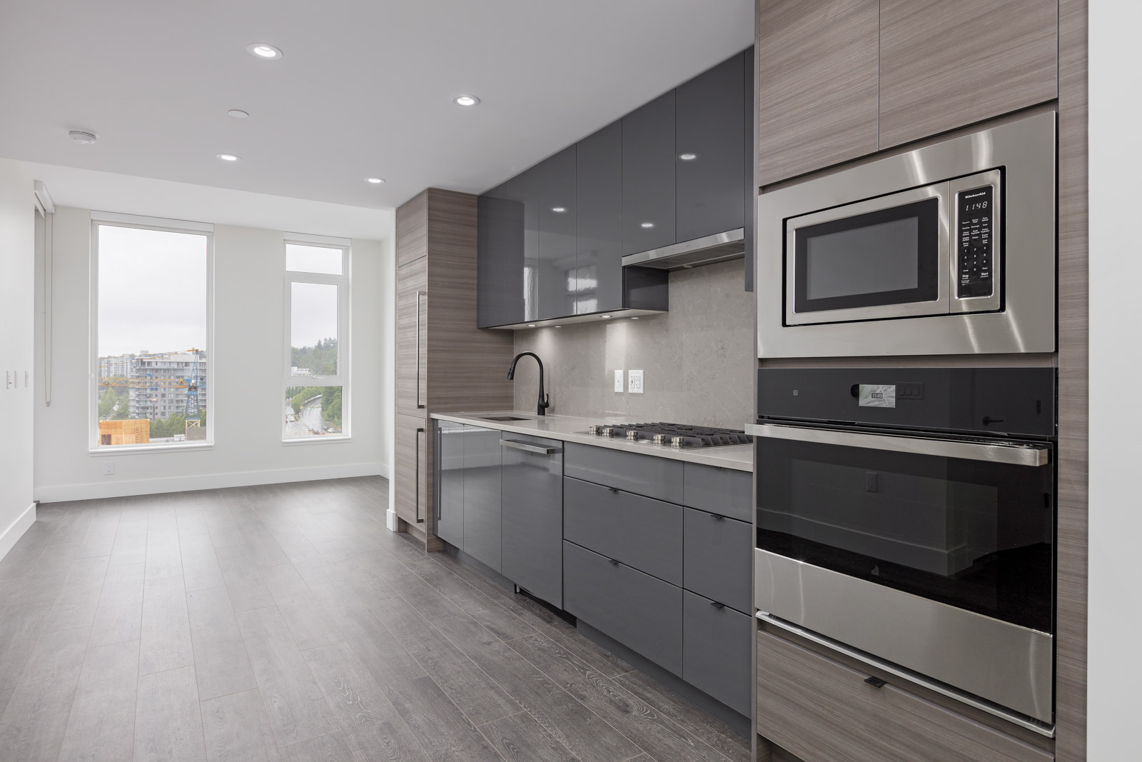 Modern kitchen with gray cabinets, built-in oven, and microwave. Large window to the left, wood flooring throughout.