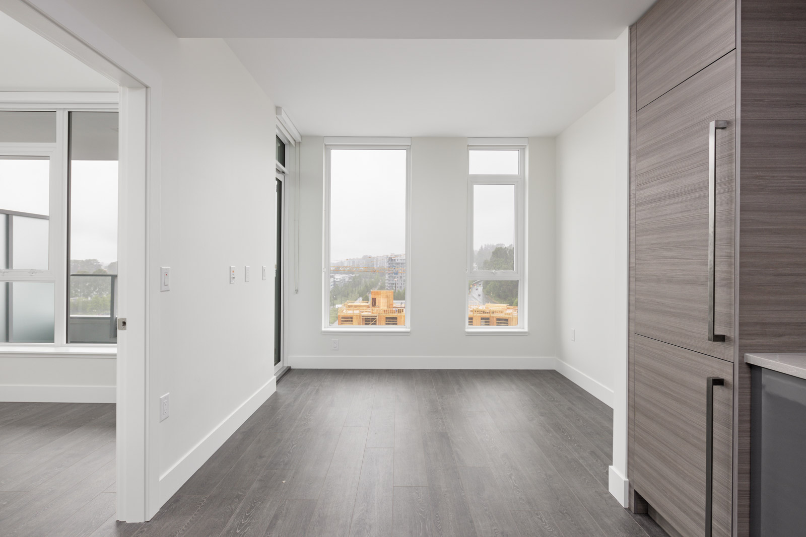 Modern, unfurnished apartment with gray hardwood floors, large windows, and a view of a balcony.