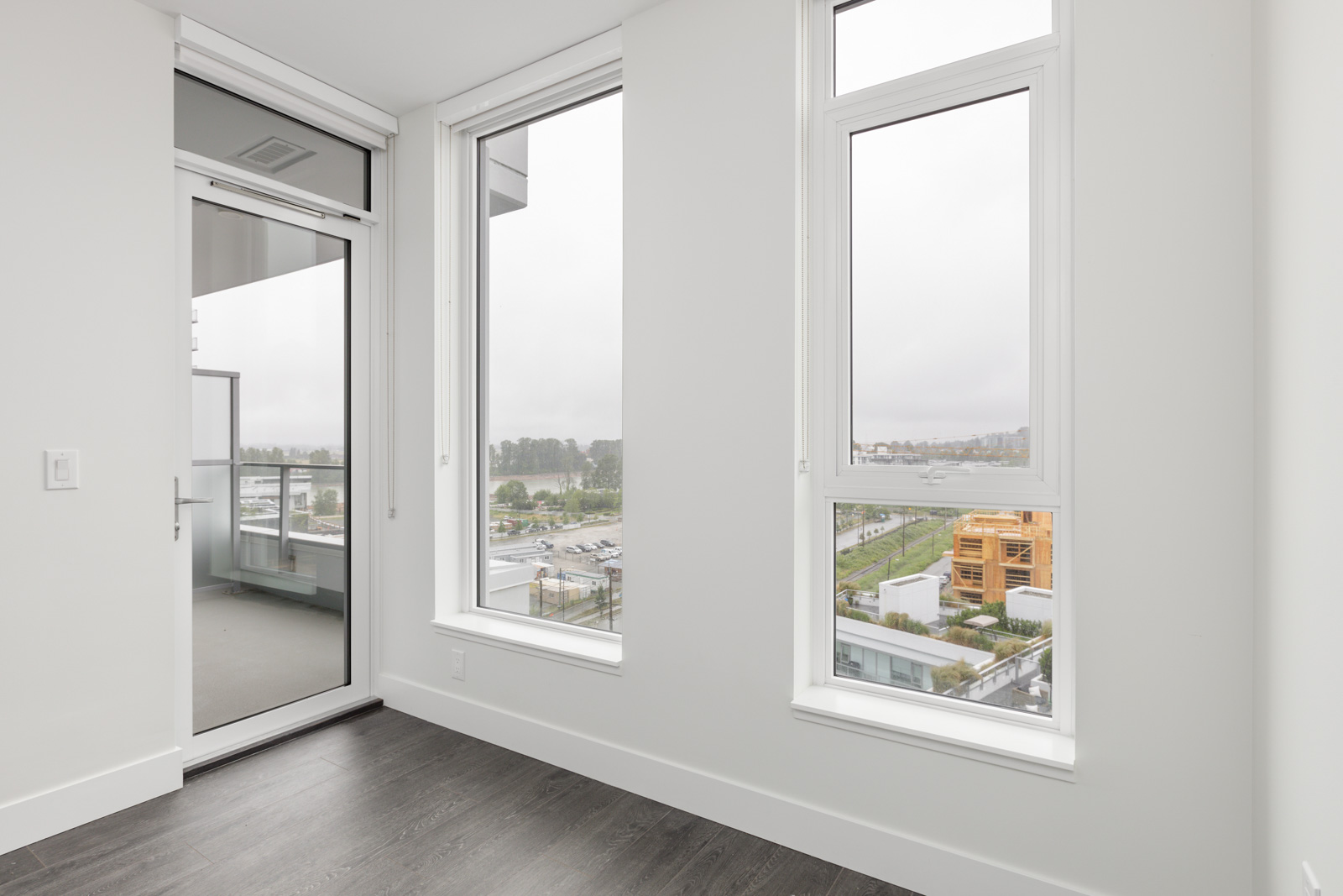 Empty room with white walls, large windows, and a glass door leading to a balcony. Dark wood flooring and a view of a cityscape through the windows.