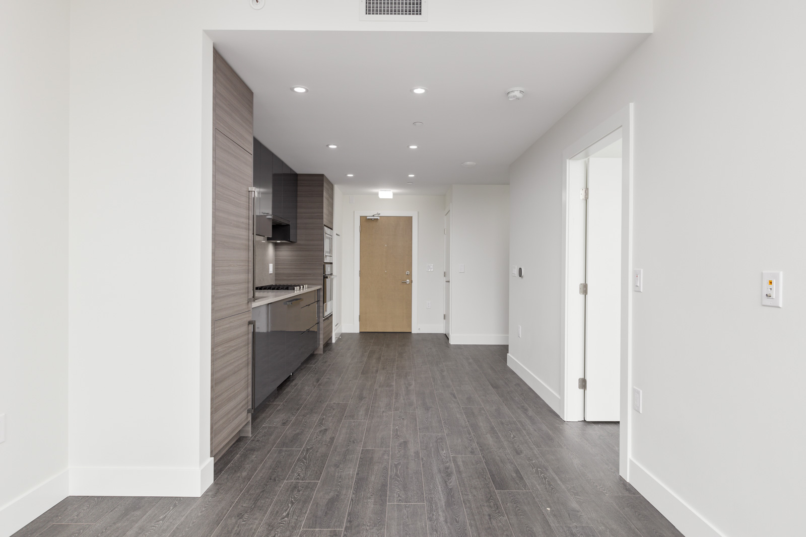 Modern, unfurnished apartment with a kitchenette, wood flooring, white walls, and recessed lighting. A brown door is visible at the end of the hallway.