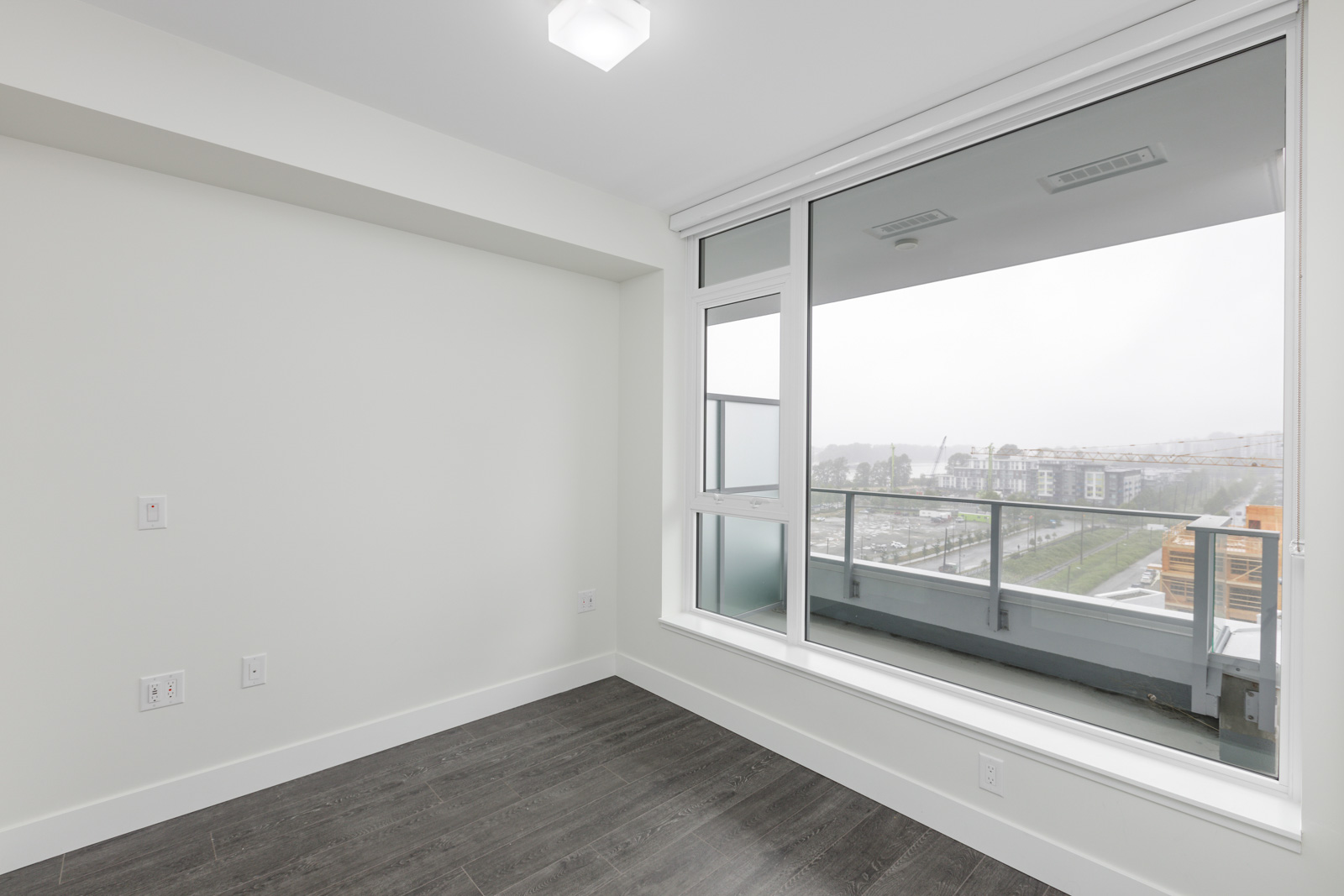 Empty room with large window and balcony view. The floor is dark wood, and the walls are light colored.