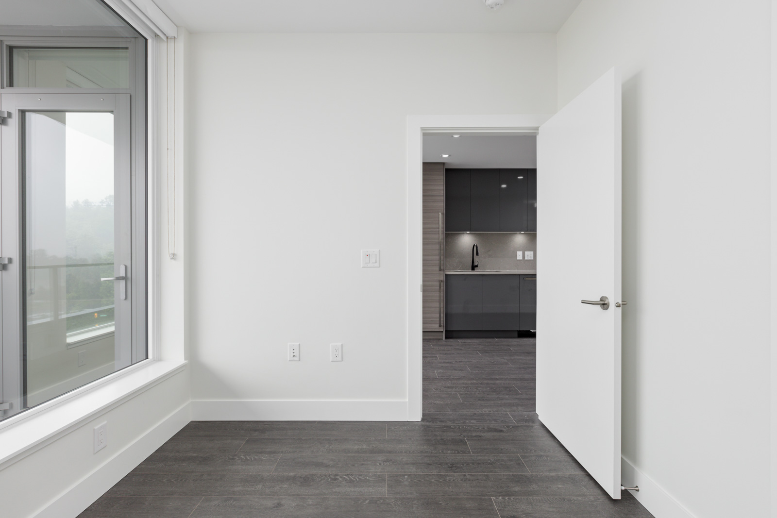Empty room with gray wooden floor and white walls, an open door leading to a kitchen with dark cabinets, and a large window on the left.
