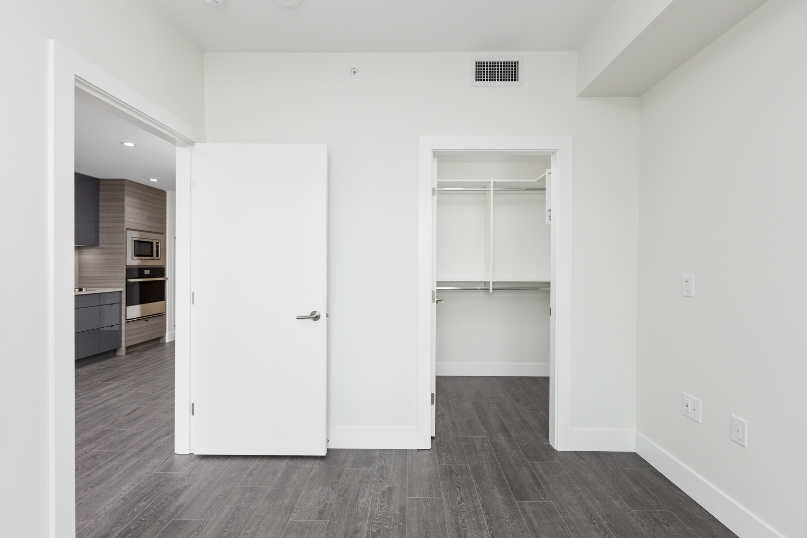 A minimalist empty room with a white door leading to a kitchen, and an open closet. The floor is dark wood, and walls are light-colored.
